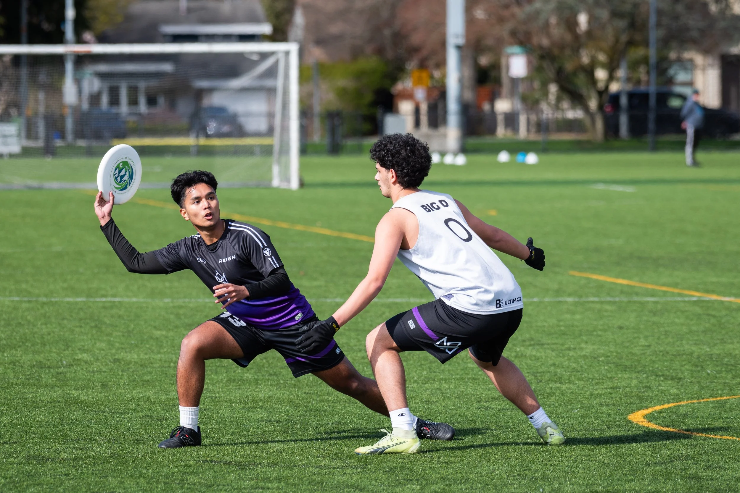 Two young men playing ultimate frisbee on a grassy field during daytime, with one about to throw the frisbee and the other attempting to block.