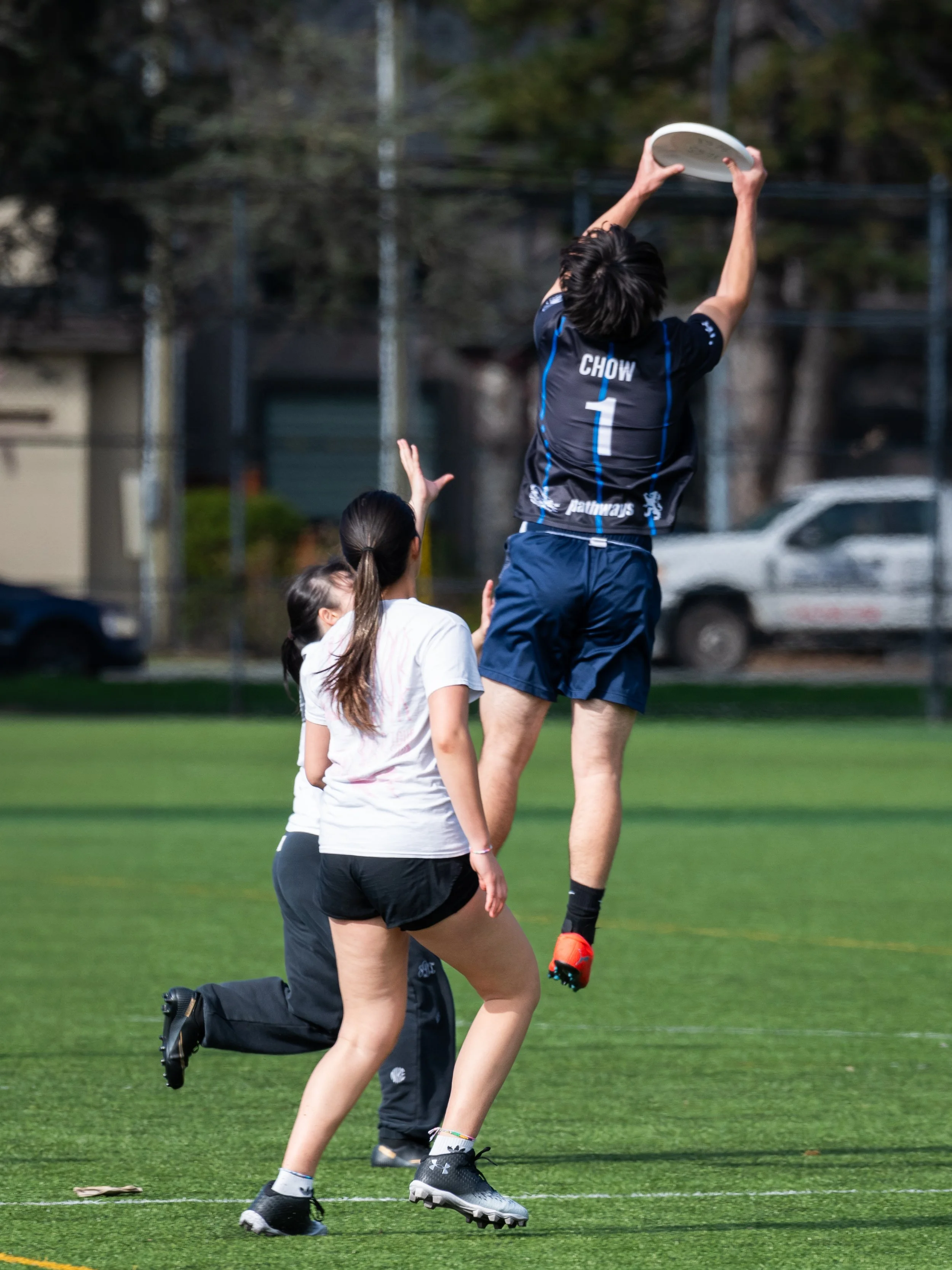 A rugby player in a blue jersey with the name 'Chow' and the number 1 jumps to catch a rugby ball during a game, with two young women in white shirts and black shorts watching and reaching up from the ground.
