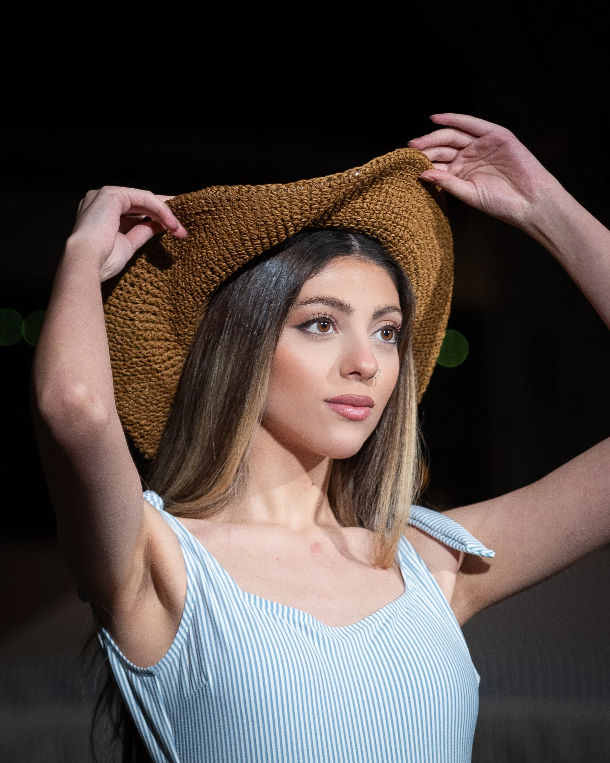 A young woman with long hair adjusting a brown wide-brimmed hat while wearing a blue and white striped sleeveless top, outdoors at night.