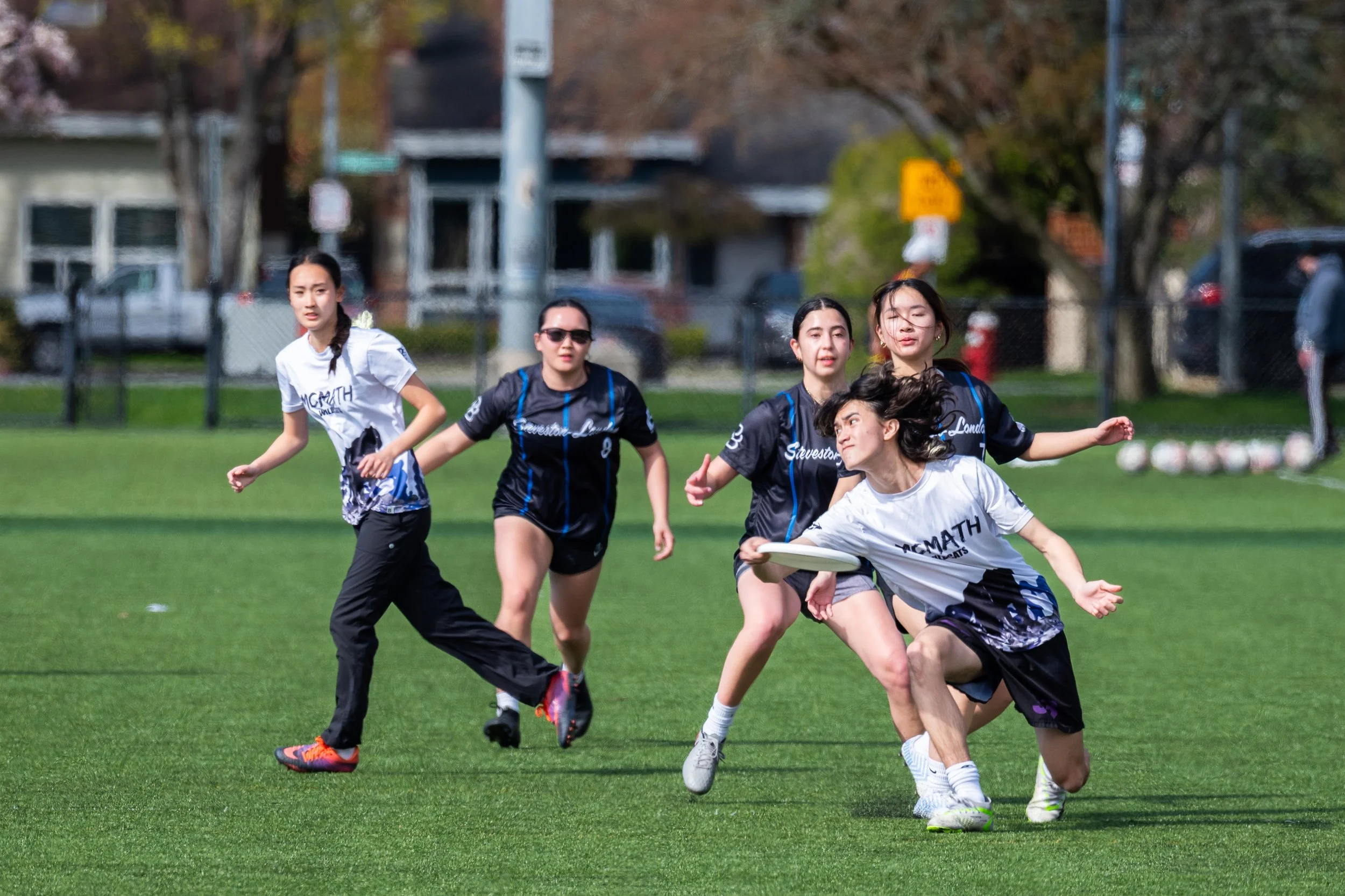 A group of women participating in a disc golf game on a grassy field, with trees and parked cars in the background.