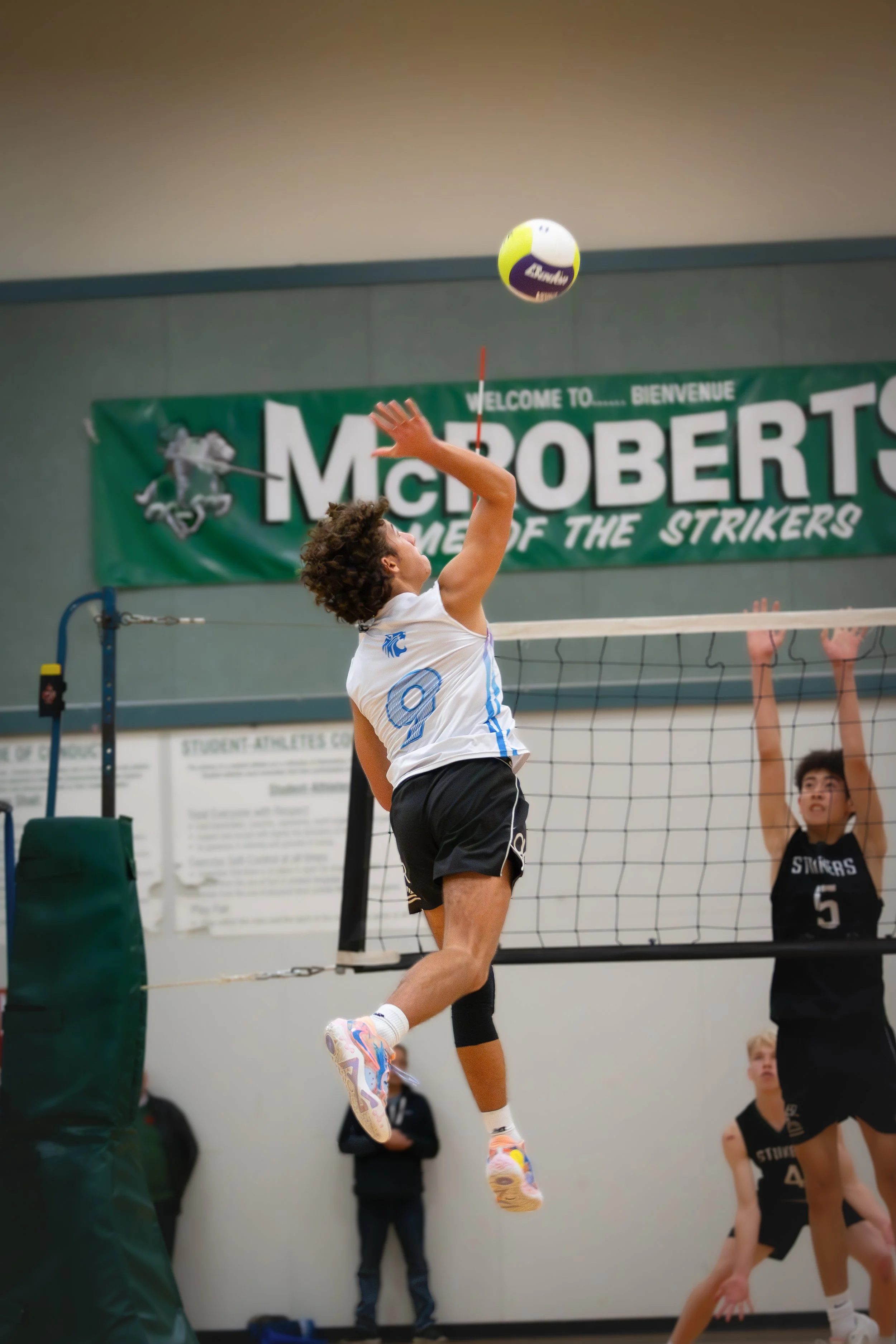 A male volleyball player wearing a white jersey with the number 9, jumping to spike the ball over the net. An opponent with a black jersey bearing the number 5 is preparing to block. The scene takes place in an indoor gym with a green banner reading 'McRoberts' in the background.