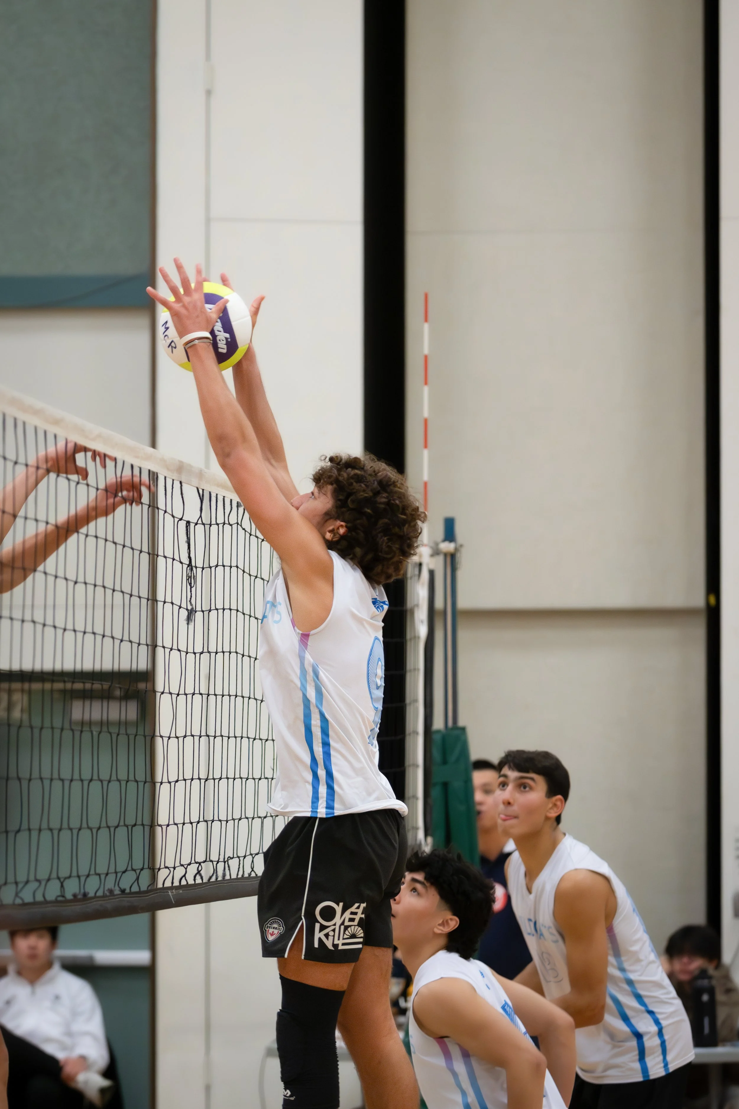 A volleyball player with curly hair jumping to block or hit the ball at the net during a game, with teammates watching. The scene takes place indoors with a volleyball net and multiple players in white jerseys.