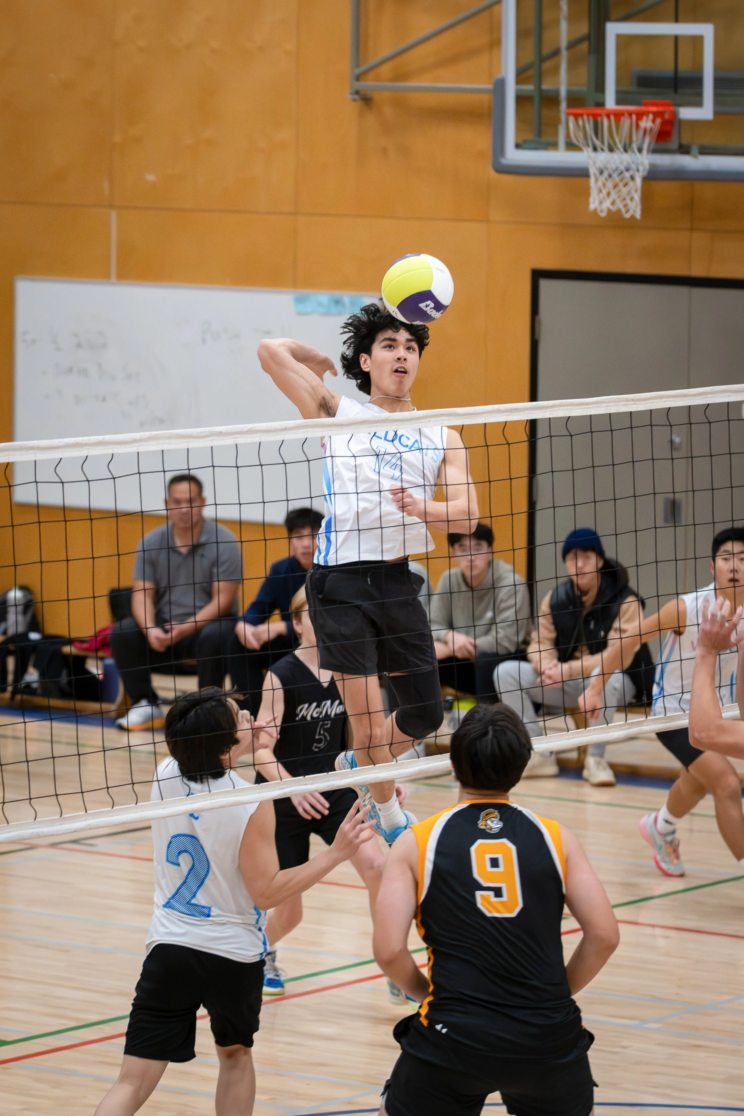 A young male volleyball player jumps at the net to spike the ball during a game in an indoor gymnasium while other players and spectators watch.
