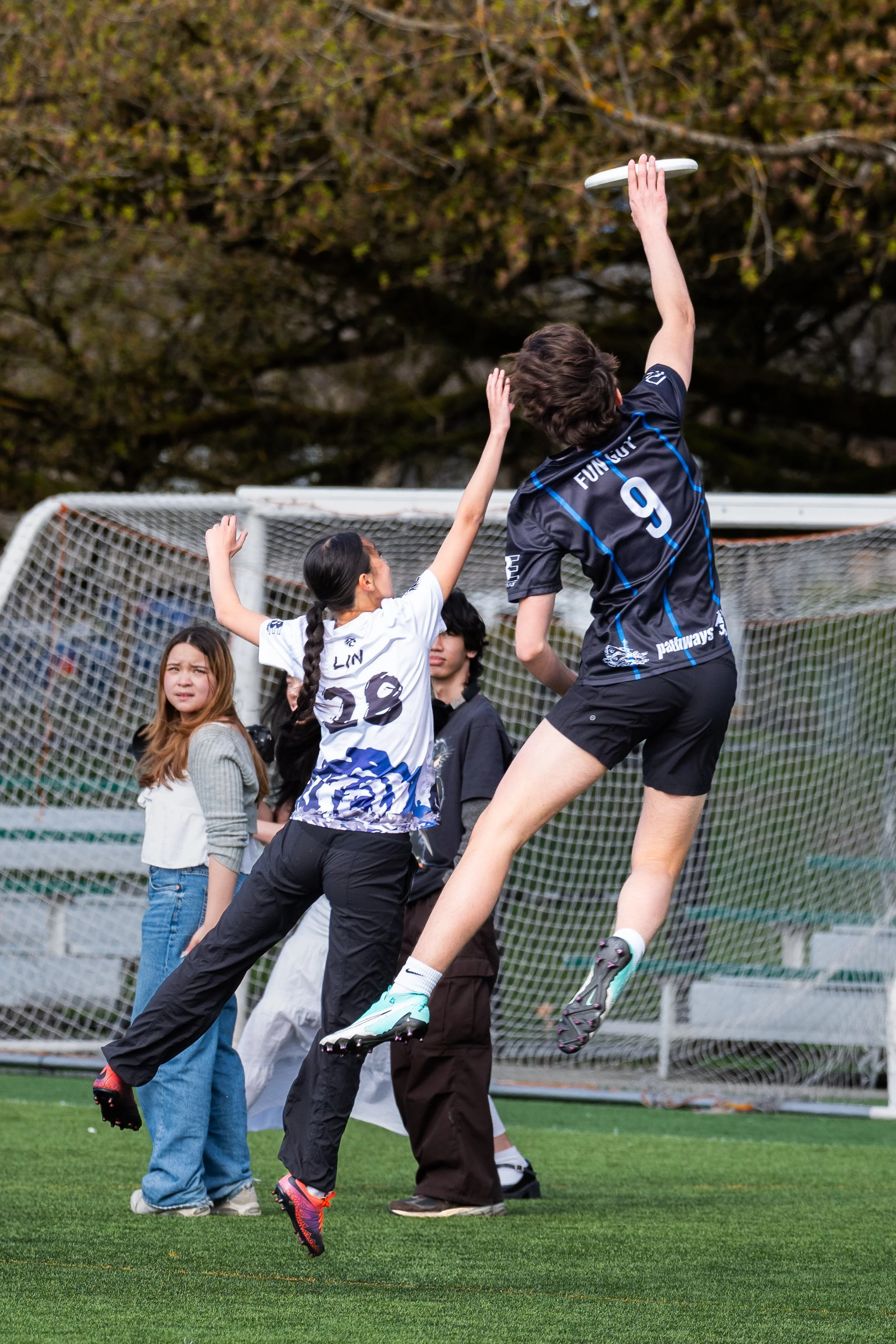 A young woman in a blue sports jersey and shorts jumps to catch a frisbee while playing soccer with a girl in a white and blue jersey on a grassy field. Two other people are nearby, with a soccer goal and net in the background.
