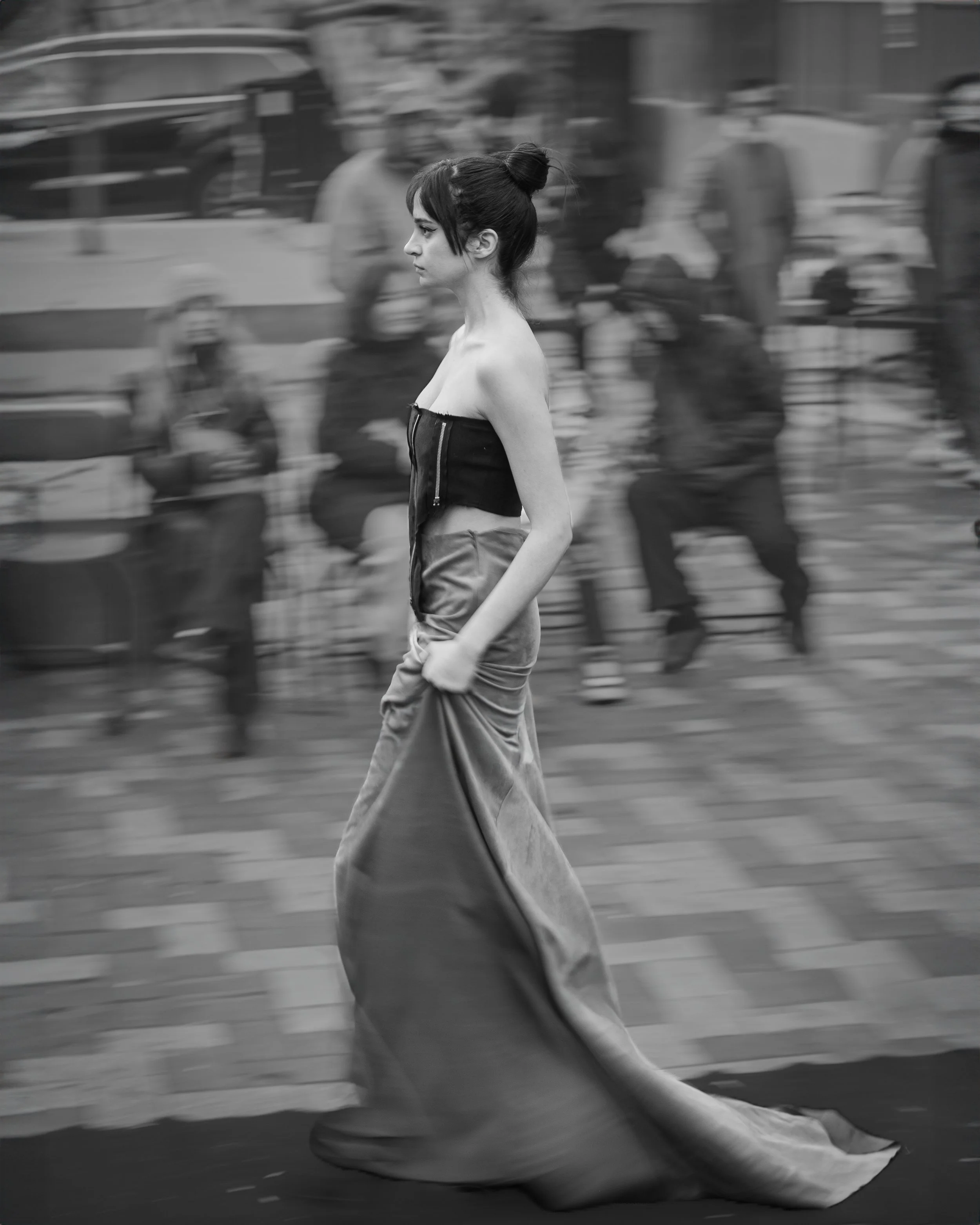 A black-and-white photo of a young woman walking in an urban environment, wearing a strapless top and a long flowing skirt, with blurred figures in the background.