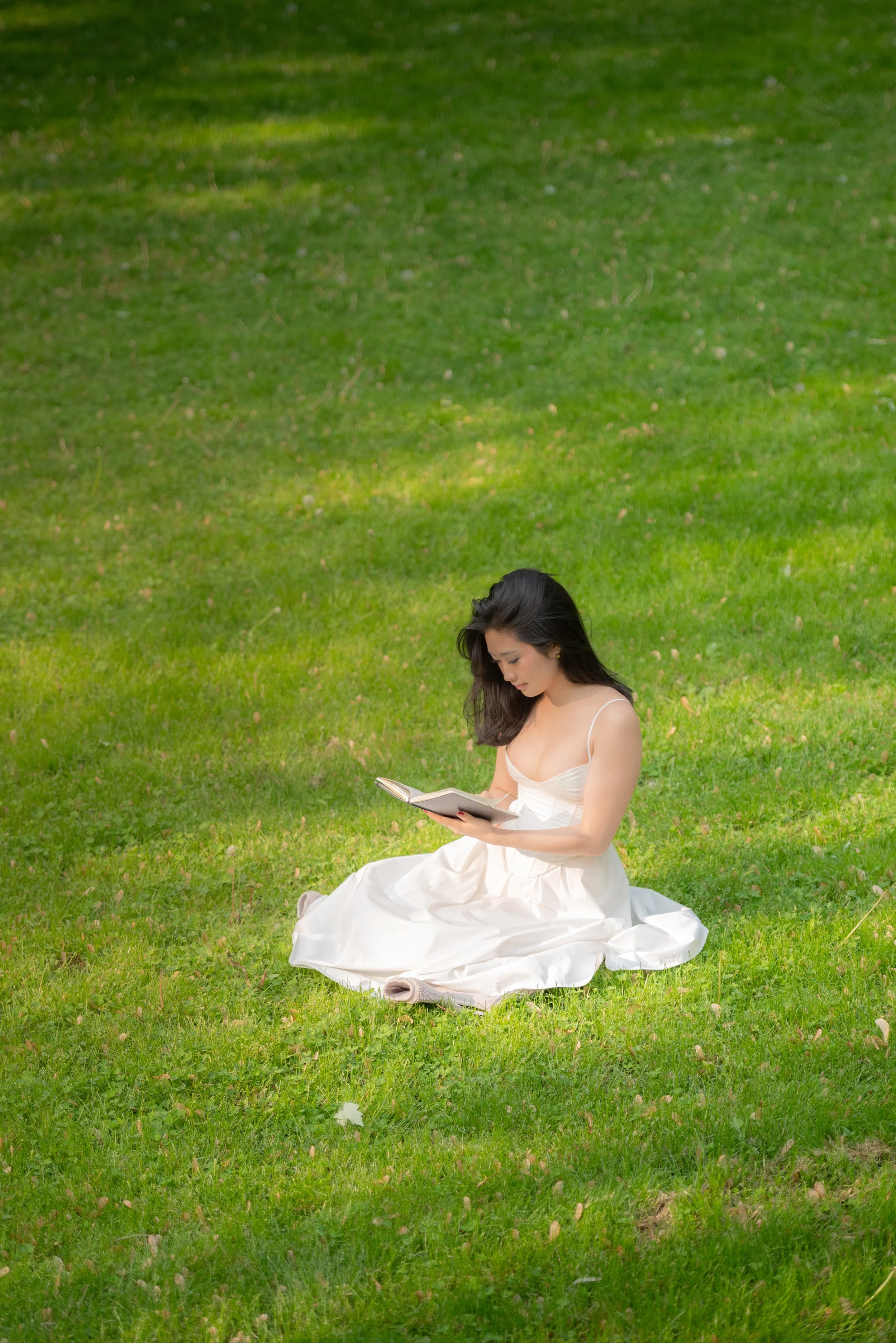 Young woman in a white dress sitting on green grass reading a book outdoors.