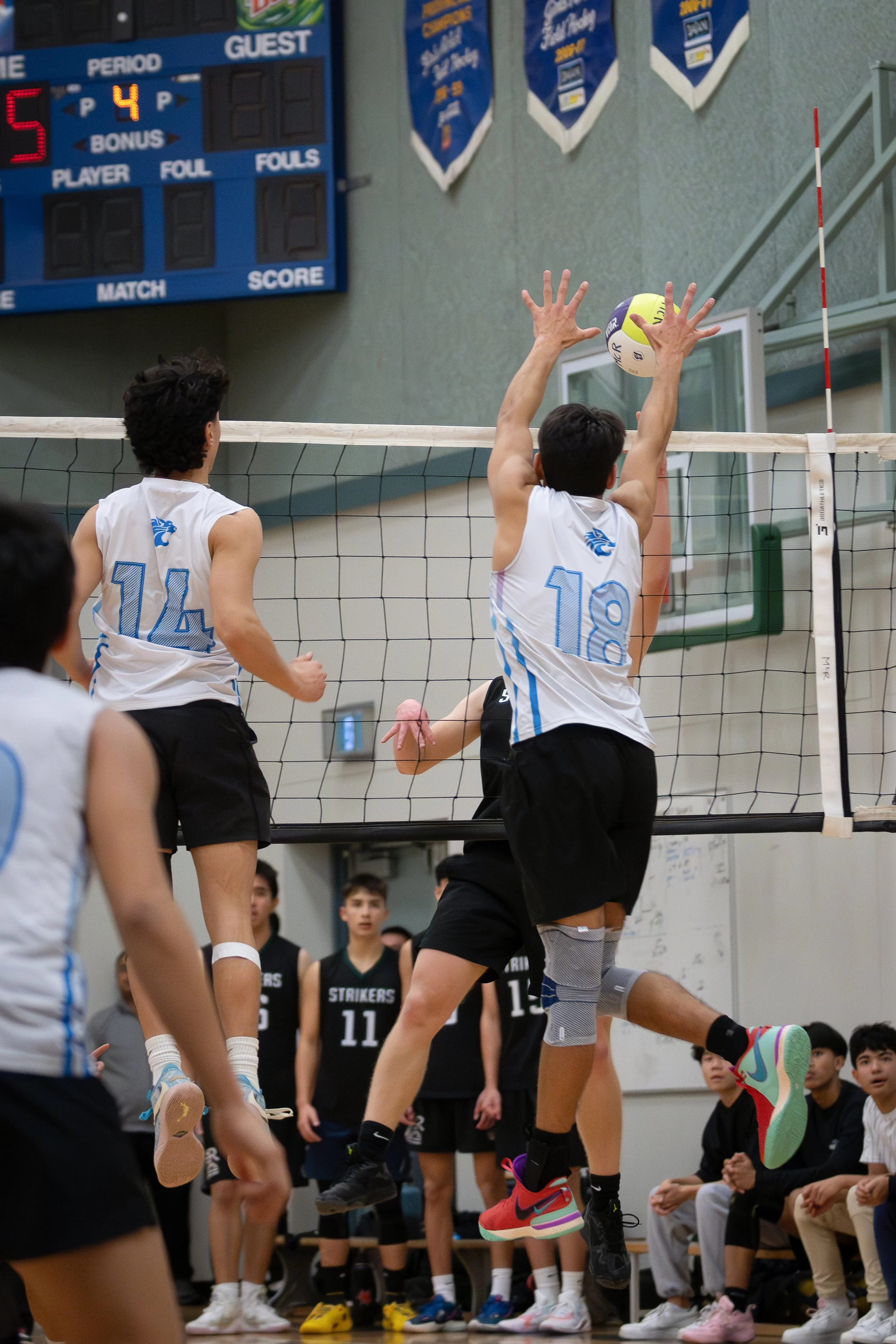 Young volleyball players competing at an indoor game, with one player jumping to spike the ball at the net while others jump to block or watch.