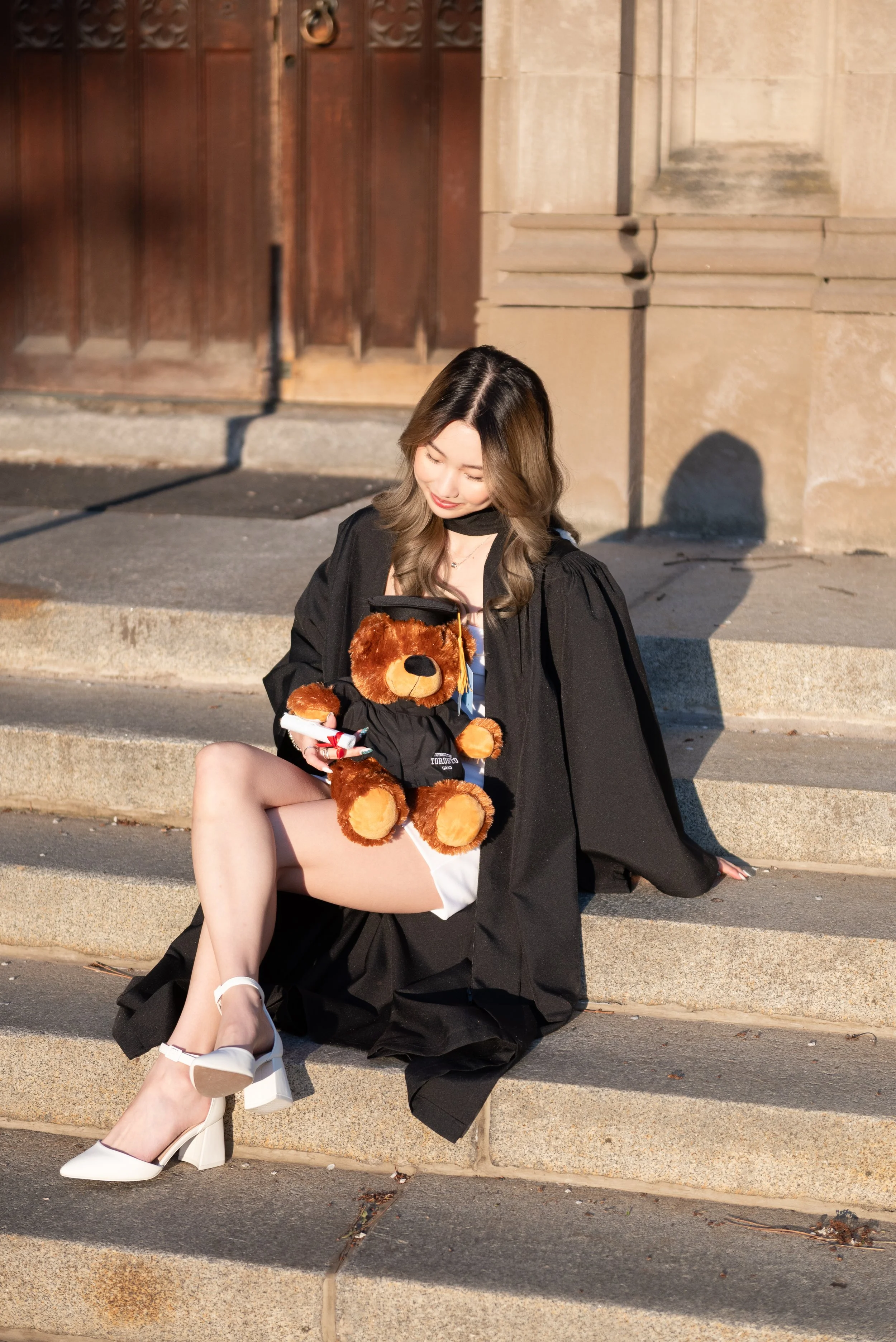 A young woman wearing a graduation gown sitting on steps outside, holding a teddy bear dressed in a graduation cap and gown, while looking at her phone.