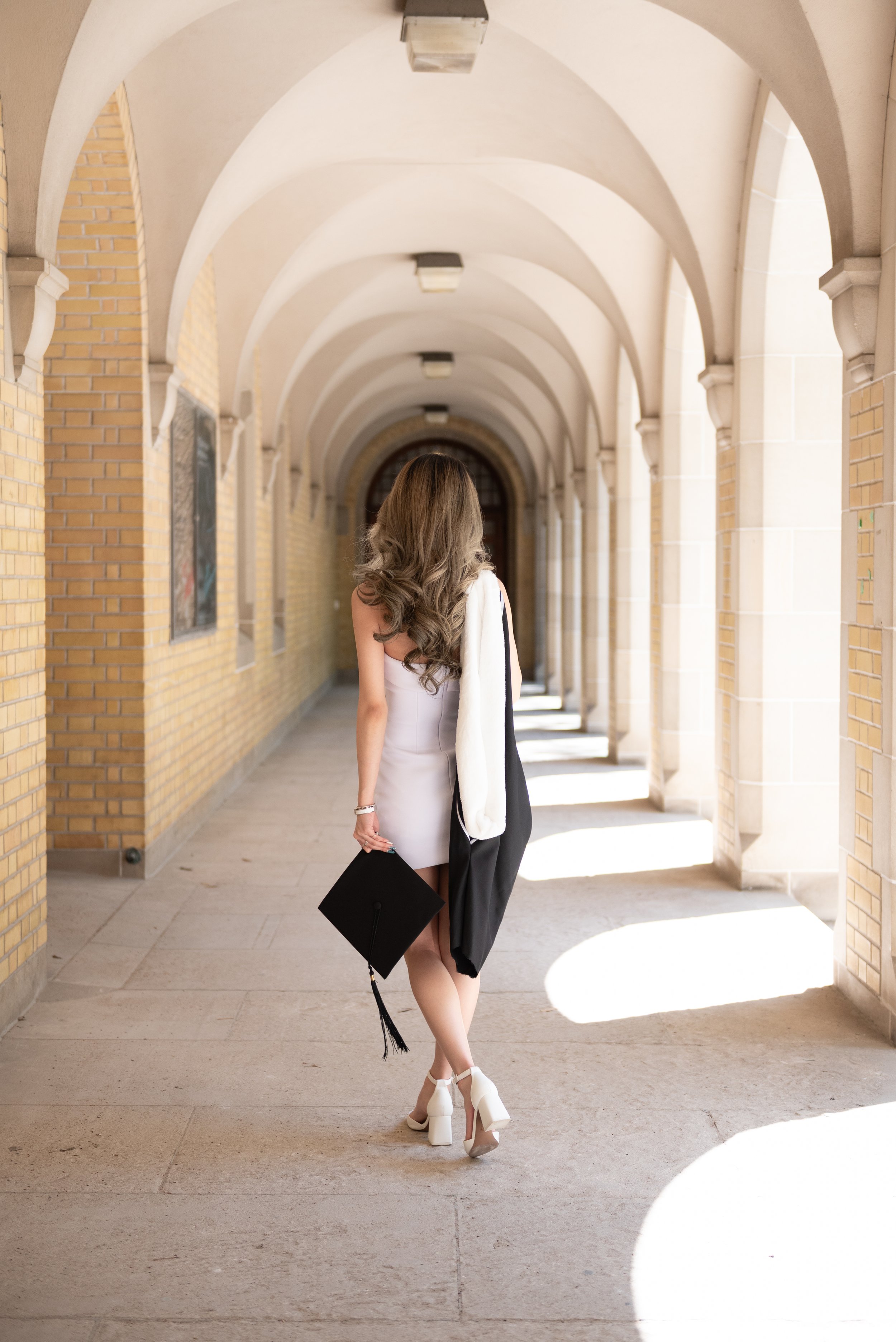 A woman with long, wavy hair in a white dress walking through a brick arched corridor, holding a black graduation cap.