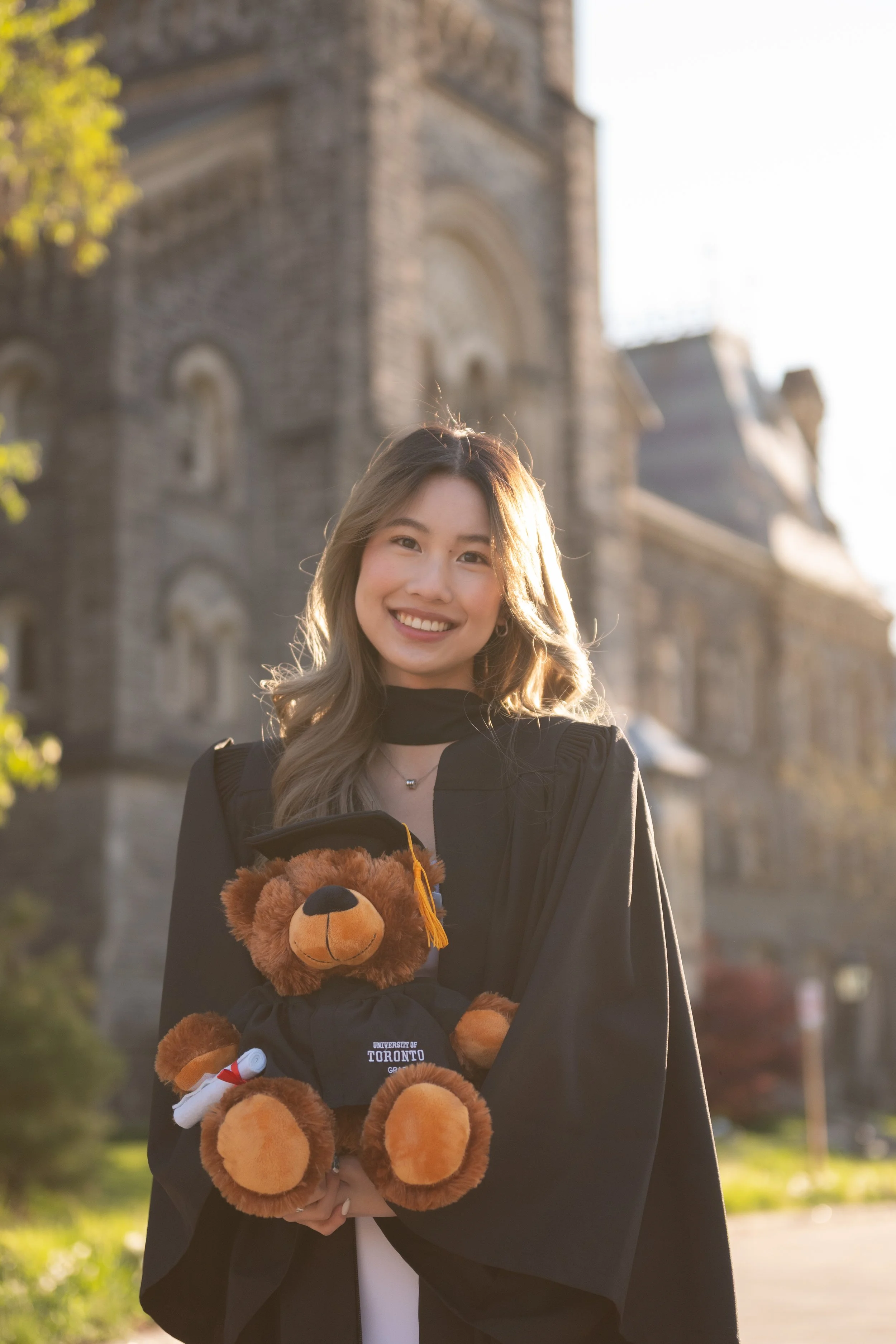 A young woman in a graduation gown holding a teddy bear with a University of Toronto cap, standing outdoors in front of a stone building.