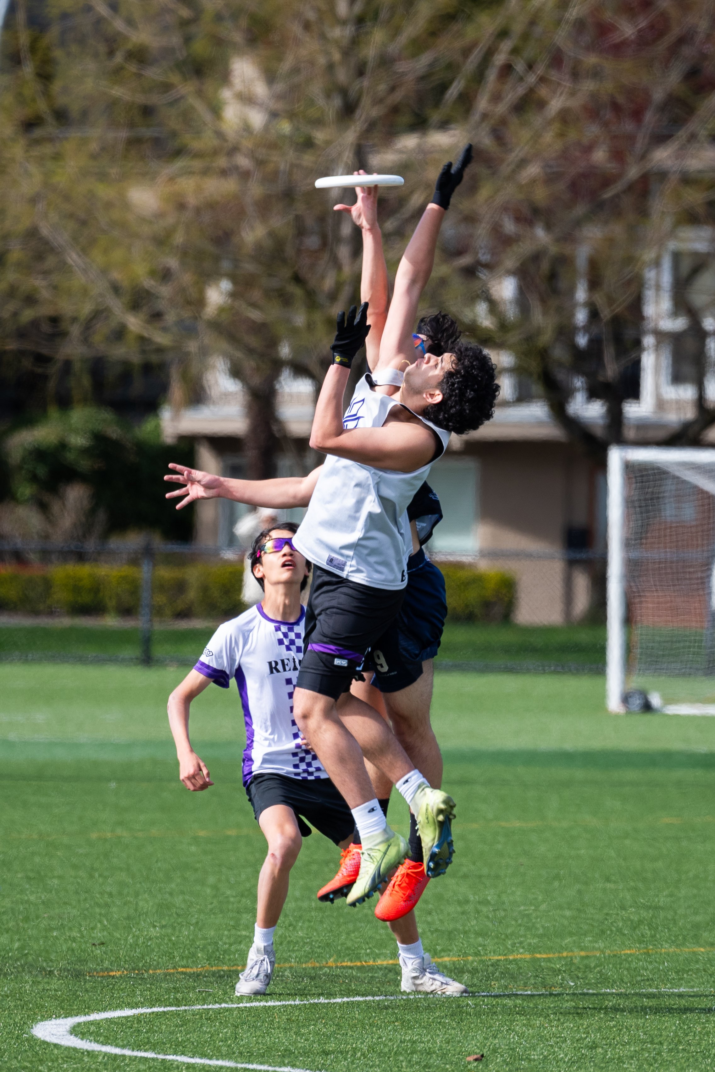 Three young athletes playing ultimate frisbee on a grassy field during daytime, with two jumping to catch the frisbee and one watching.
