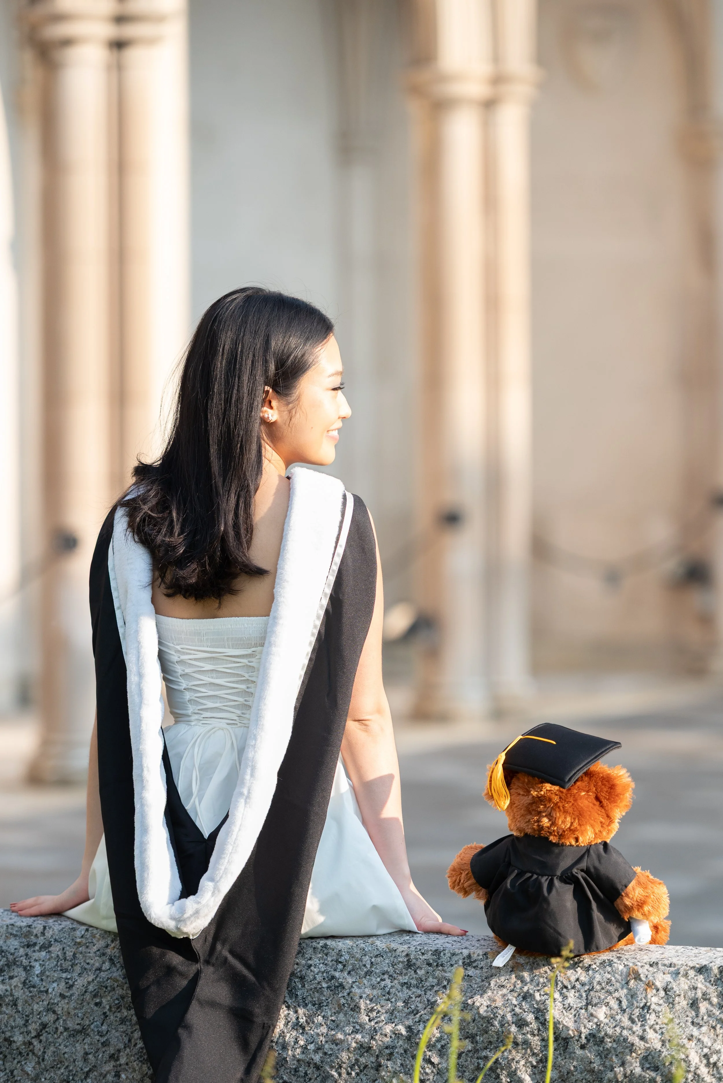 A young woman in a white dress with a black graduation stole, sitting on a stone ledge, smiling and looking away. Next to her is a teddy bear dressed in a cap and gown, also sitting on the ledge, with a background of large columns.