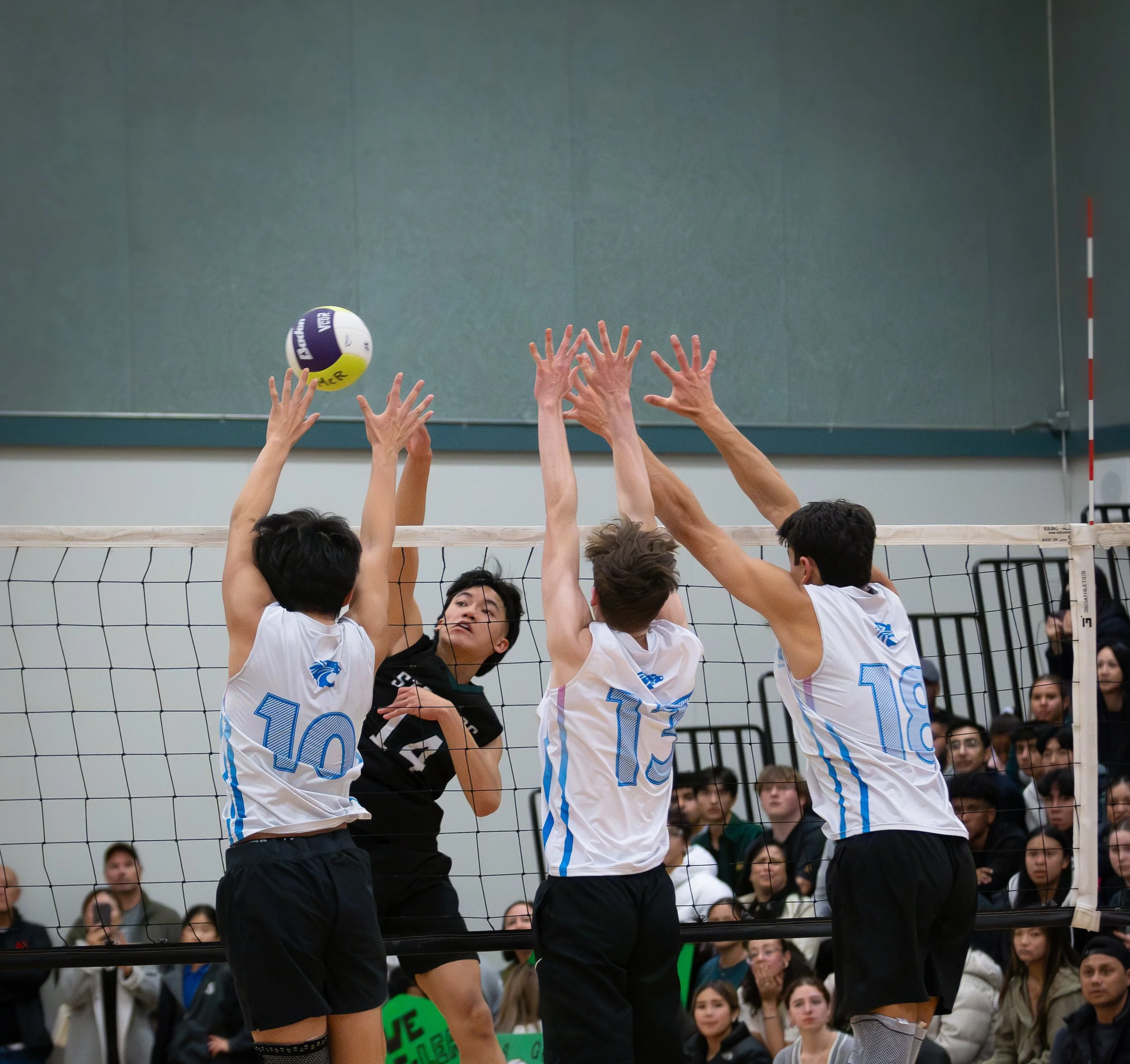 Four volleyball players jumping to block a volleyball at the net during a match with an audience watching in the background.