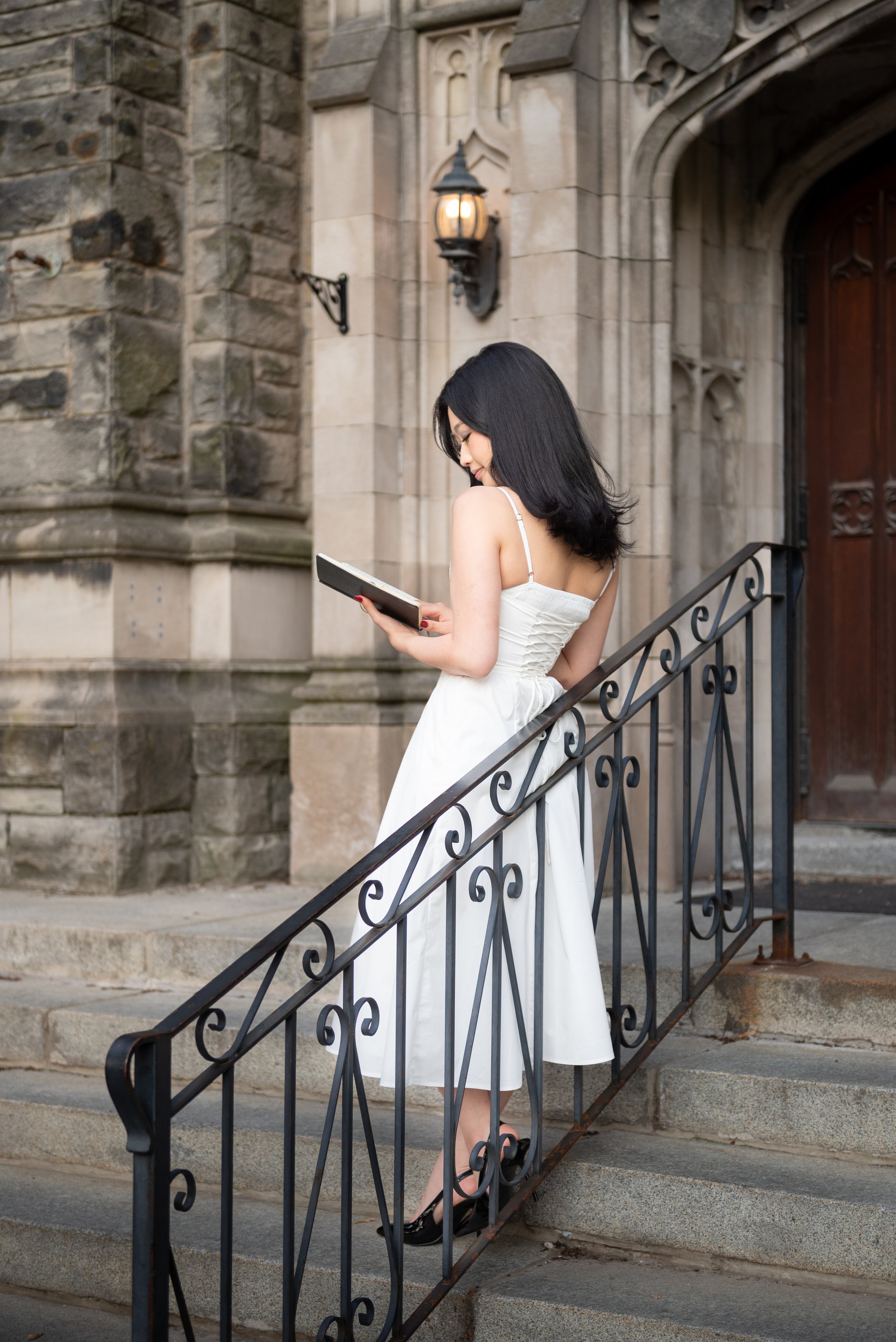 A young woman in a white dress and black heels standing on the steps of a stone building, reading a book.