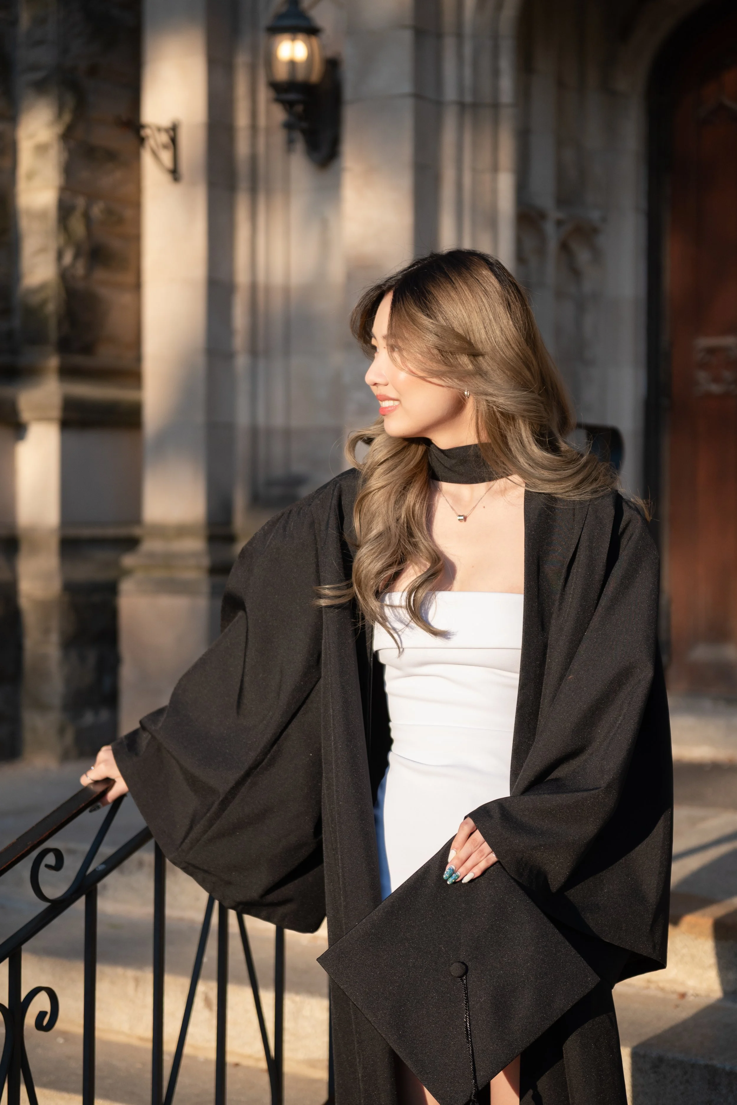 A young woman wearing a graduation gown holding a diploma, standing outside a historic stone building during sunset.