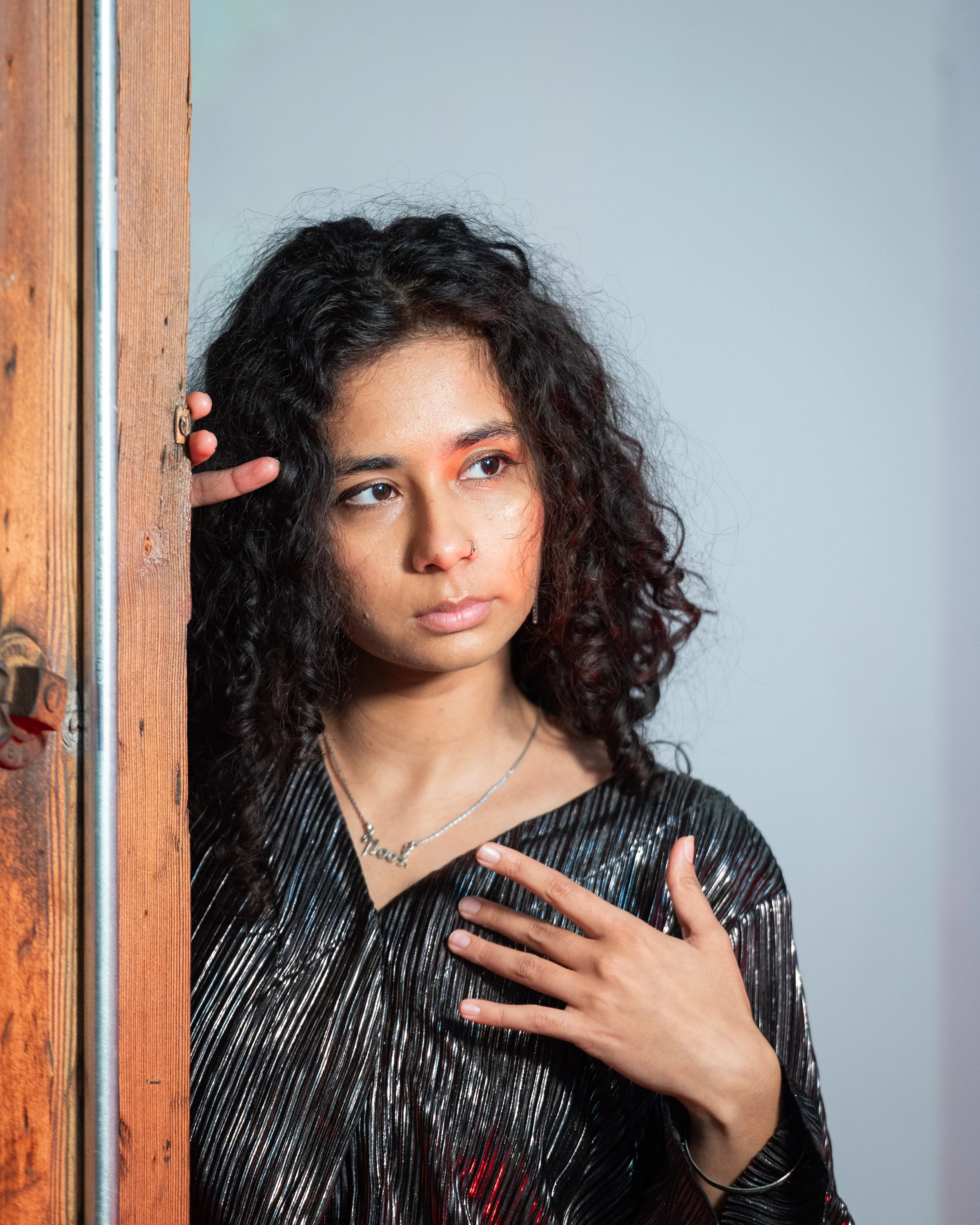 A young woman with curly dark hair and a nose piercing wearing a black shiny dress, looking thoughtfully to the side, partially hiding behind a wooden door.