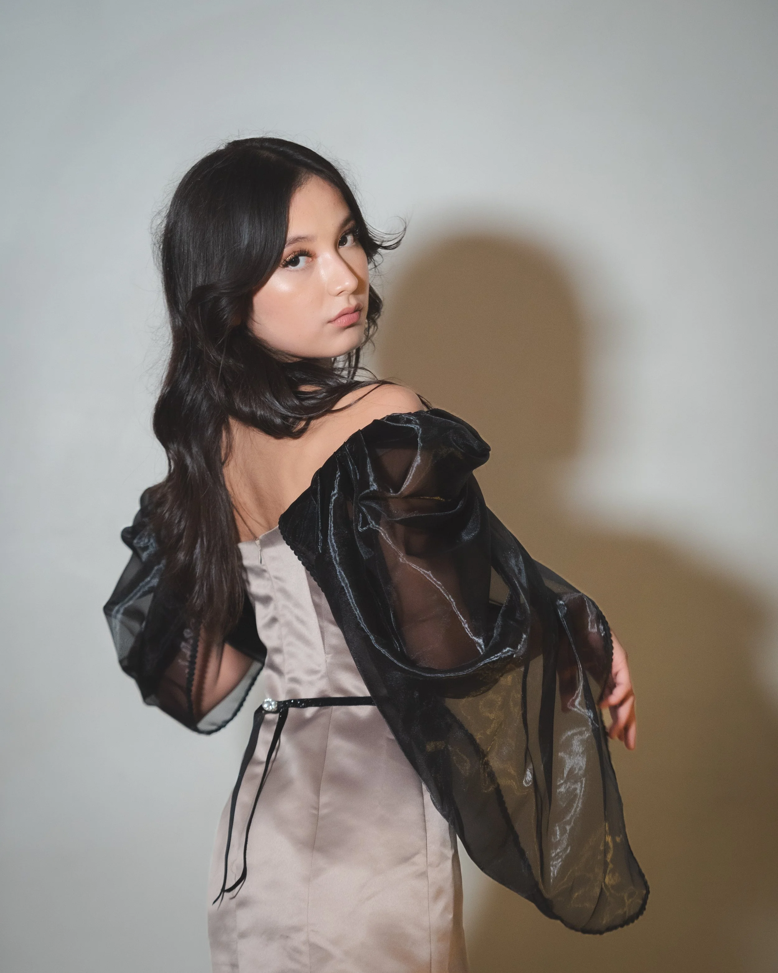 A young woman with long dark hair wearing a shiny beige dress with black sheer puff sleeves, standing against a plain wall, looking over her shoulder.
