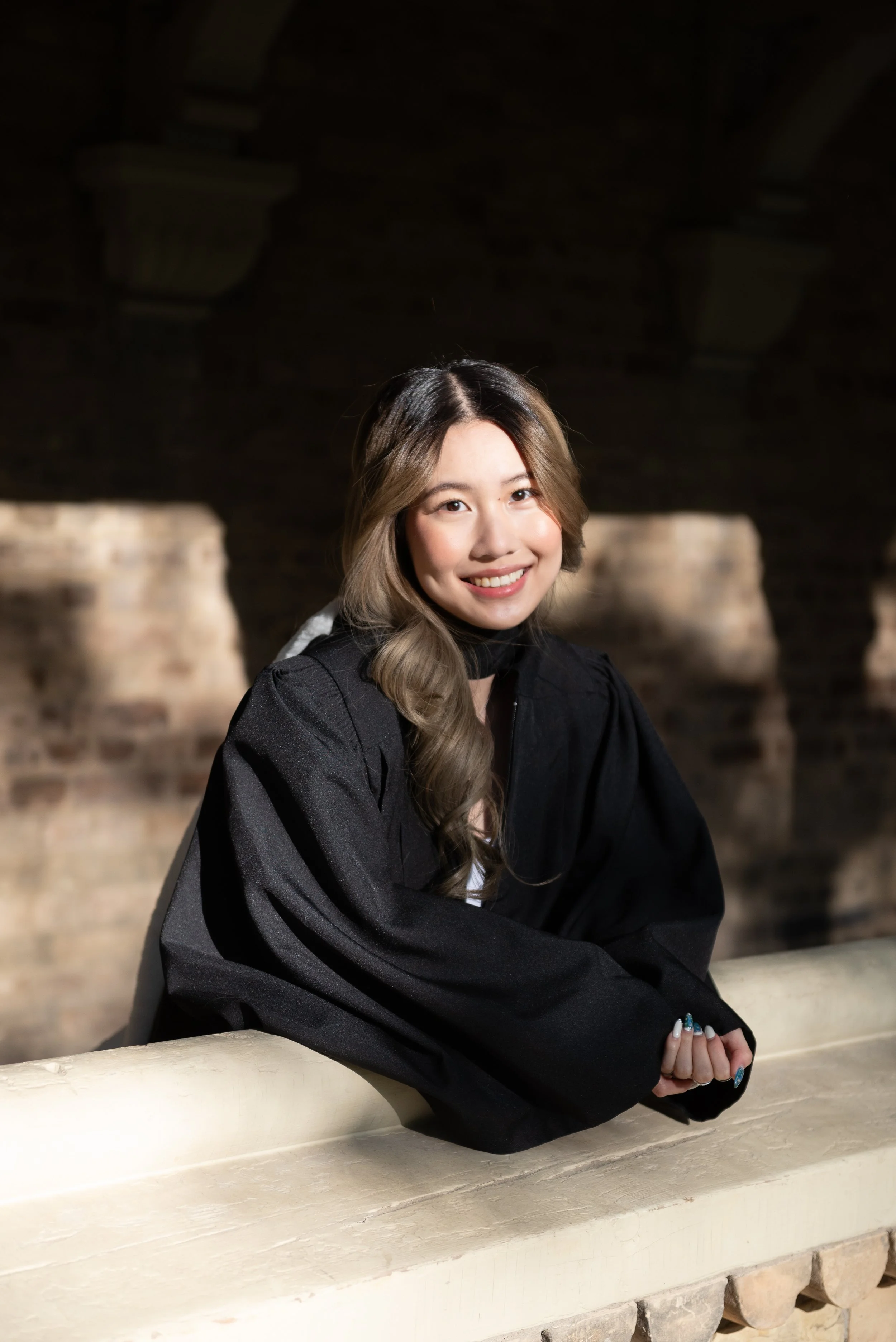 A young woman in a black graduation gown smiling while leaning on a railing inside a brick building.