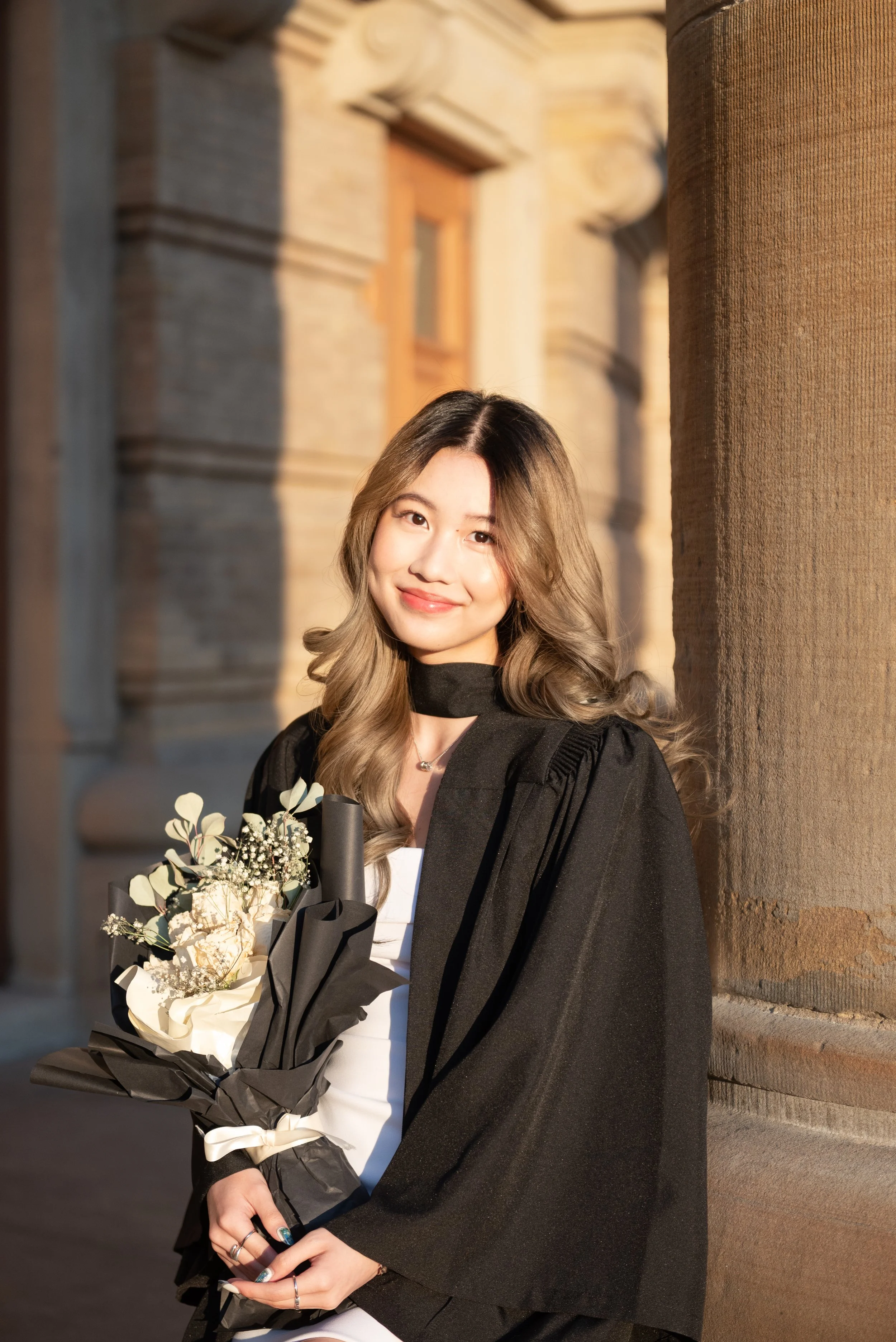 Young woman in graduation gown holding a bouquet of flowers, standing outdoors beside a stone column, smiling at the camera during sunset.