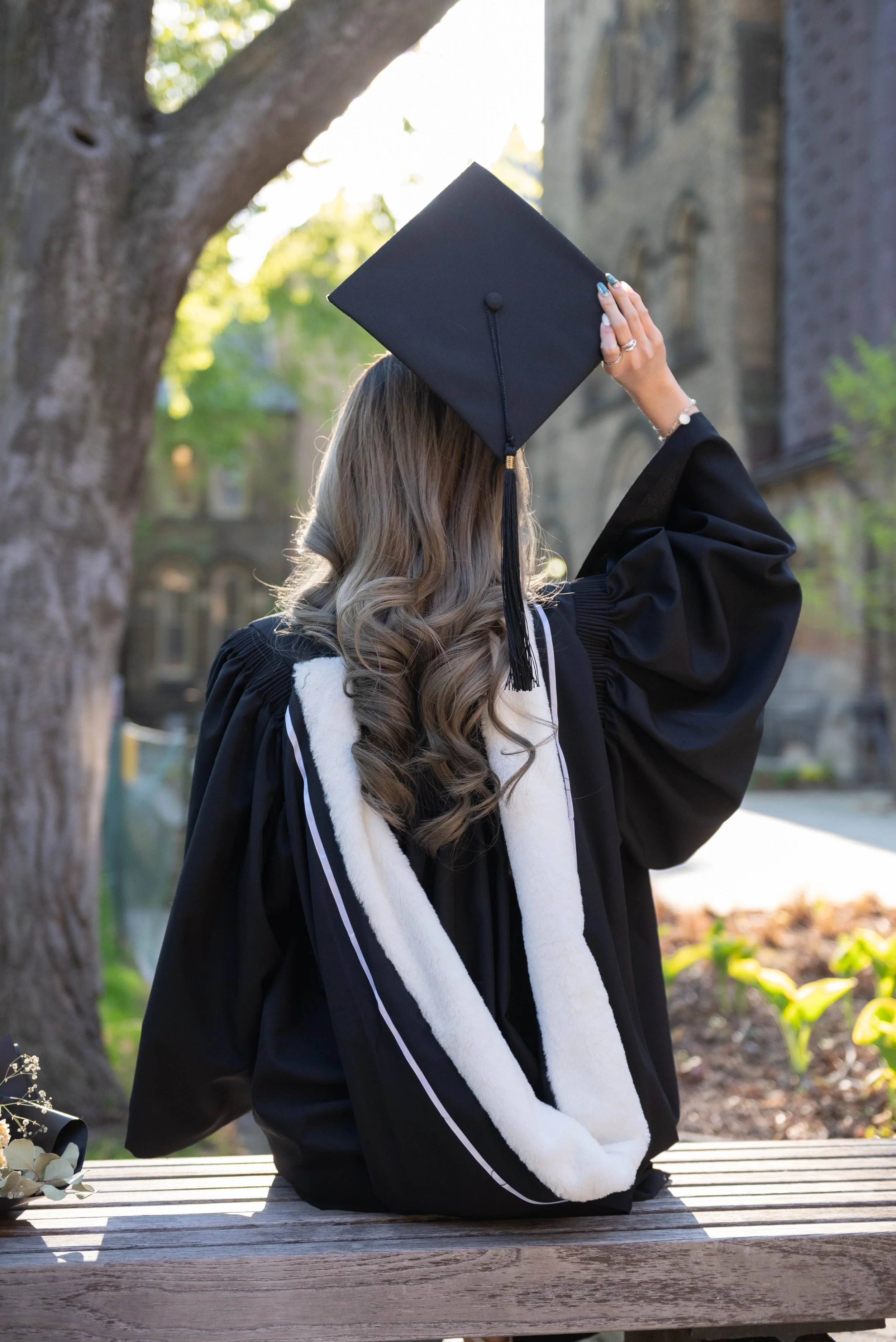 A woman wearing a graduation gown and cap, sitting on a bench outdoors, with her back toward the camera.