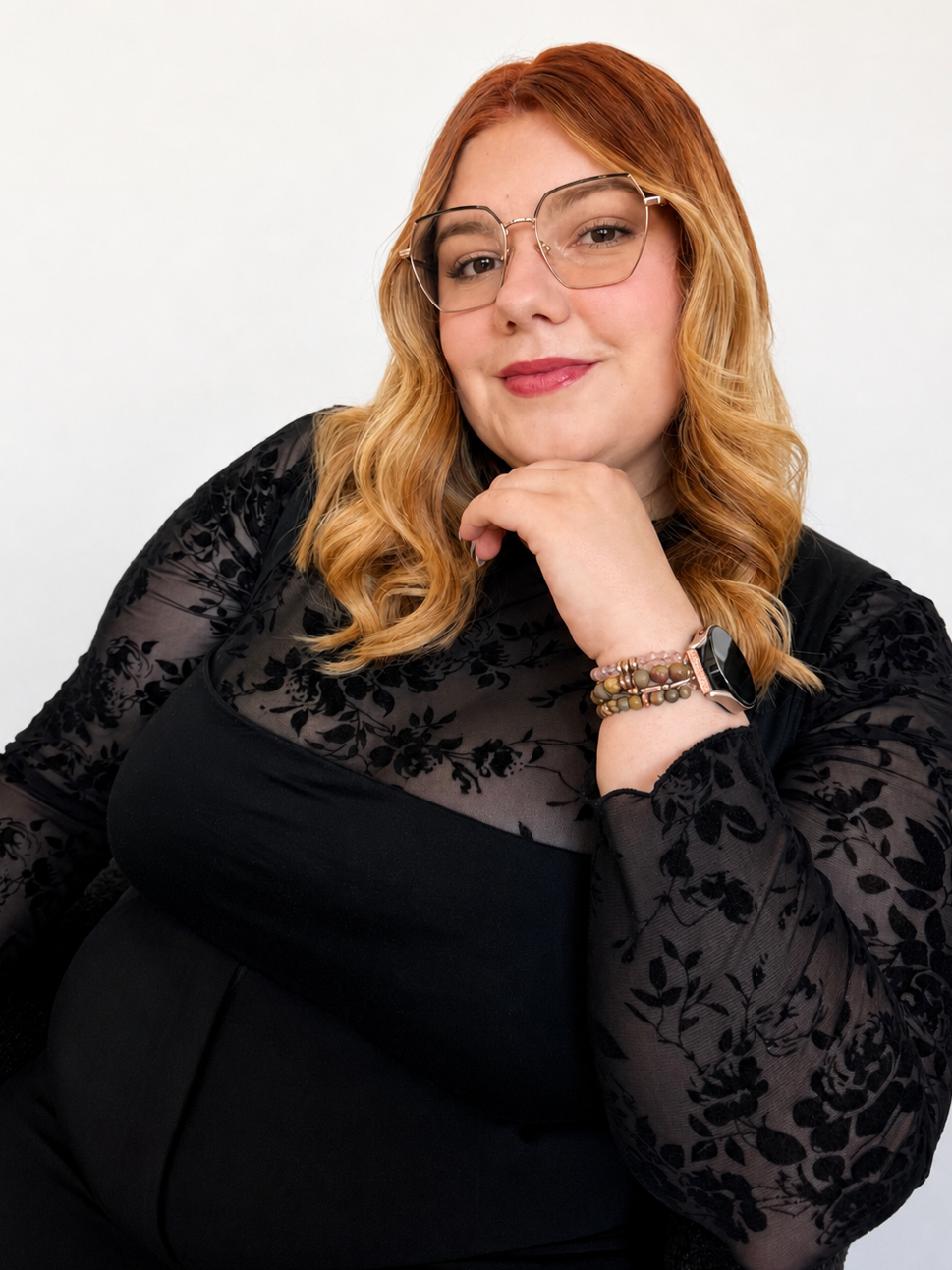 A woman with red hair wearing glasses, a black lace top with floral patterns, and a wristwatch, posing against a plain white background.