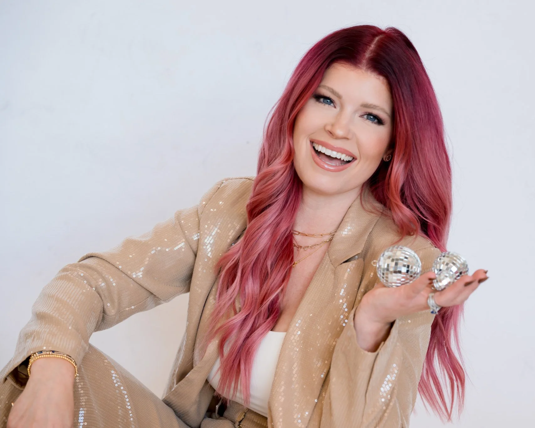 A smiling woman with long, wavy pink hair holding two small disco balls, wearing a beige sequin blazer and multiple gold necklaces, against a plain light background.