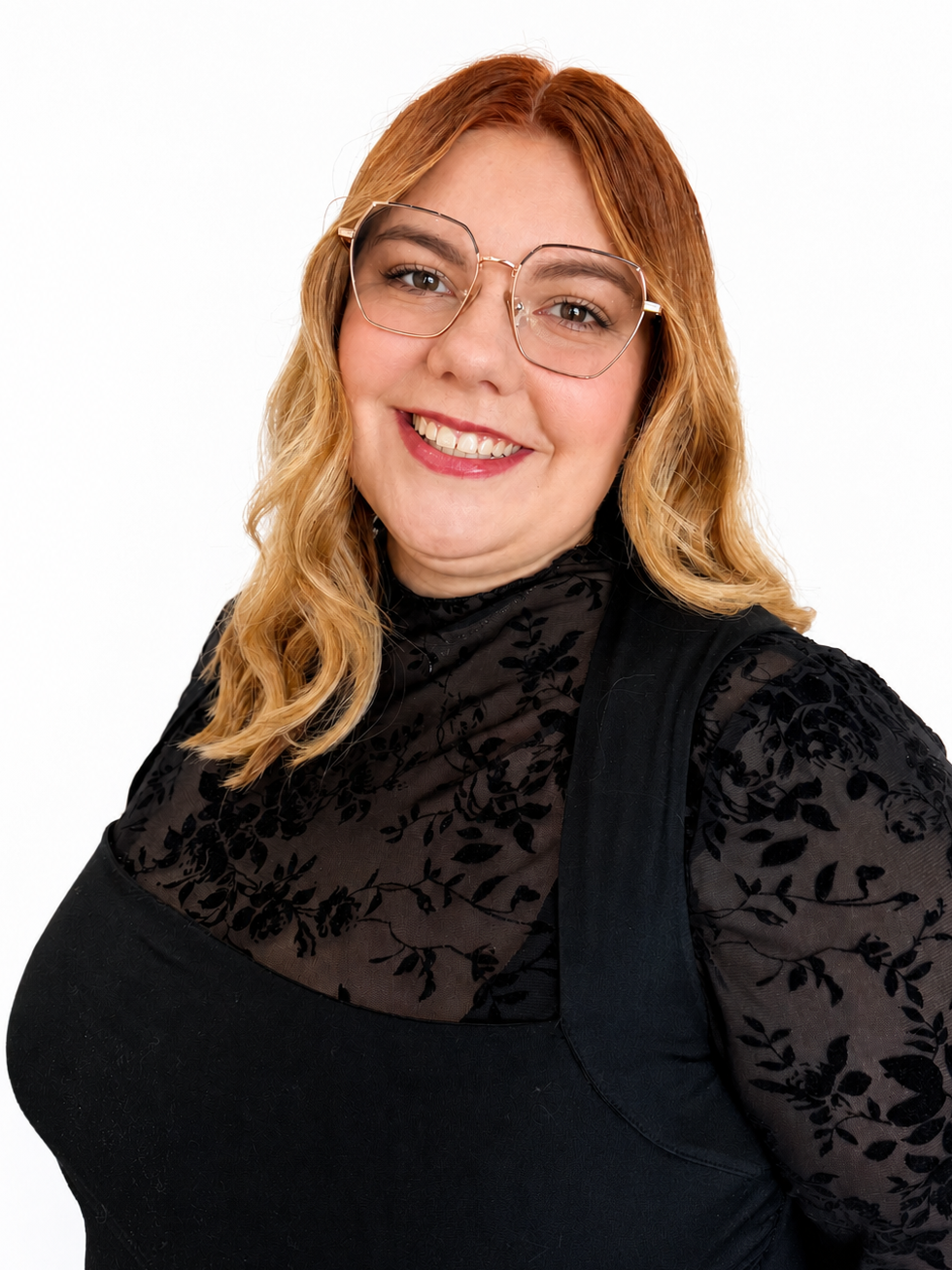A woman with red hair wearing glasses, a black, floral-patterned sheer top, and a black vest, smiling at the camera against a white background.