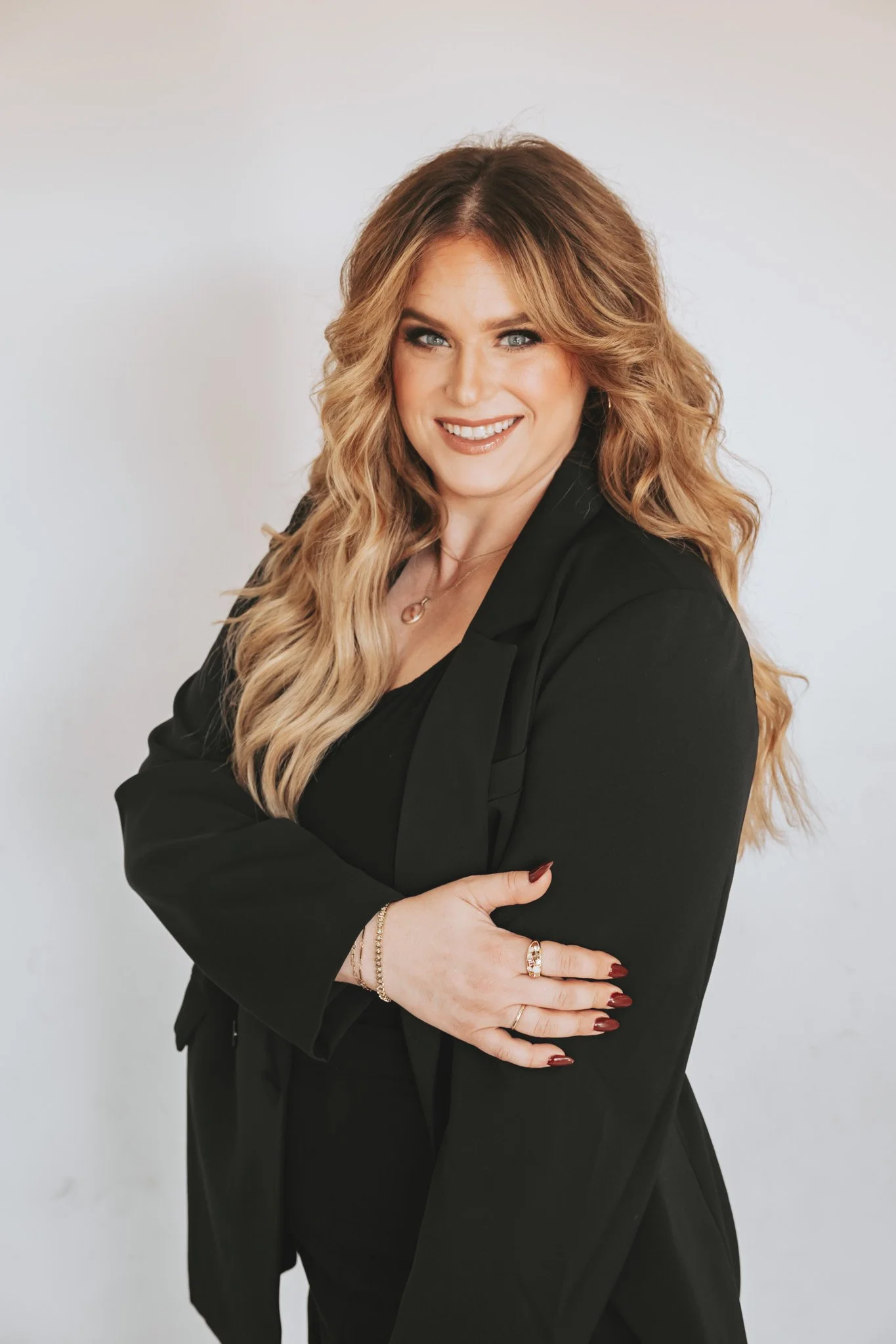 A woman smiling outdoors wearing a wide-brimmed tan hat, gold jewelry, and a blue floral dress.
