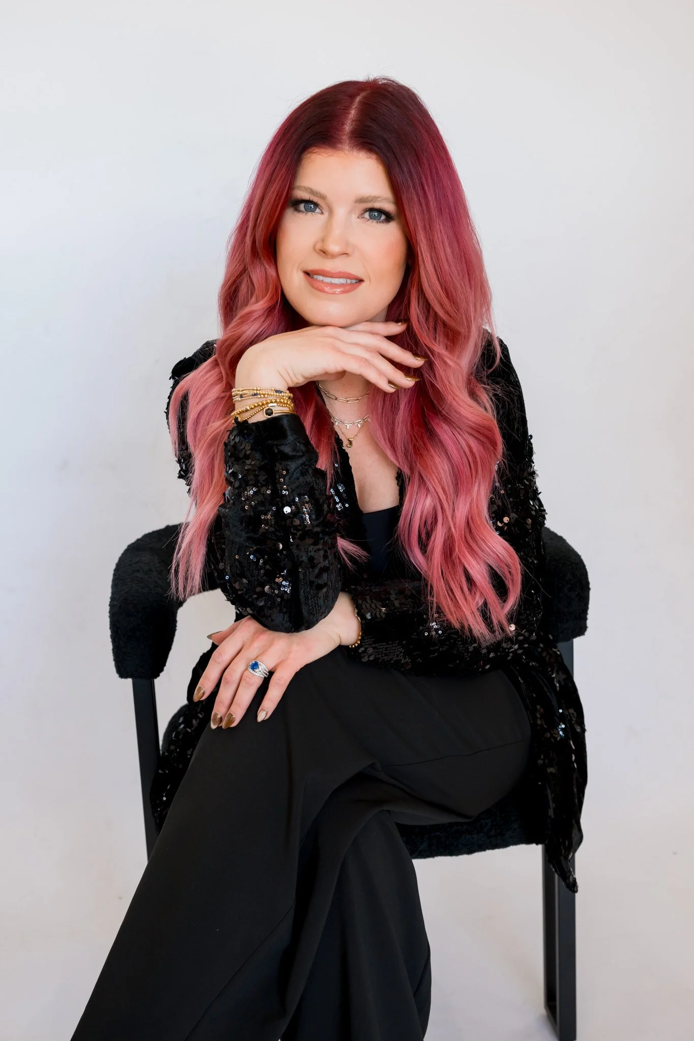 A woman with vibrant red and pink hair, smiling and looking to the side, wearing a black sequin blazer with long sleeves.