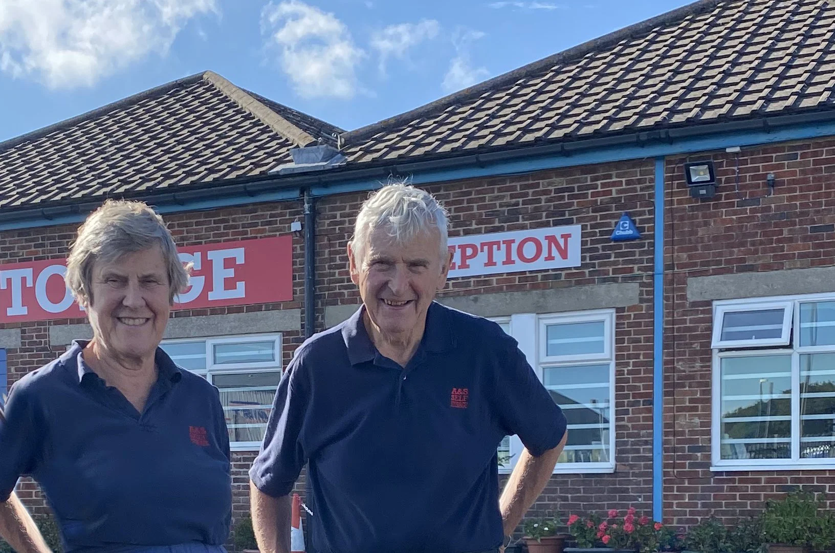 Two older men standing outside a building with a sign that says 'A&S Self Storage Reception'.