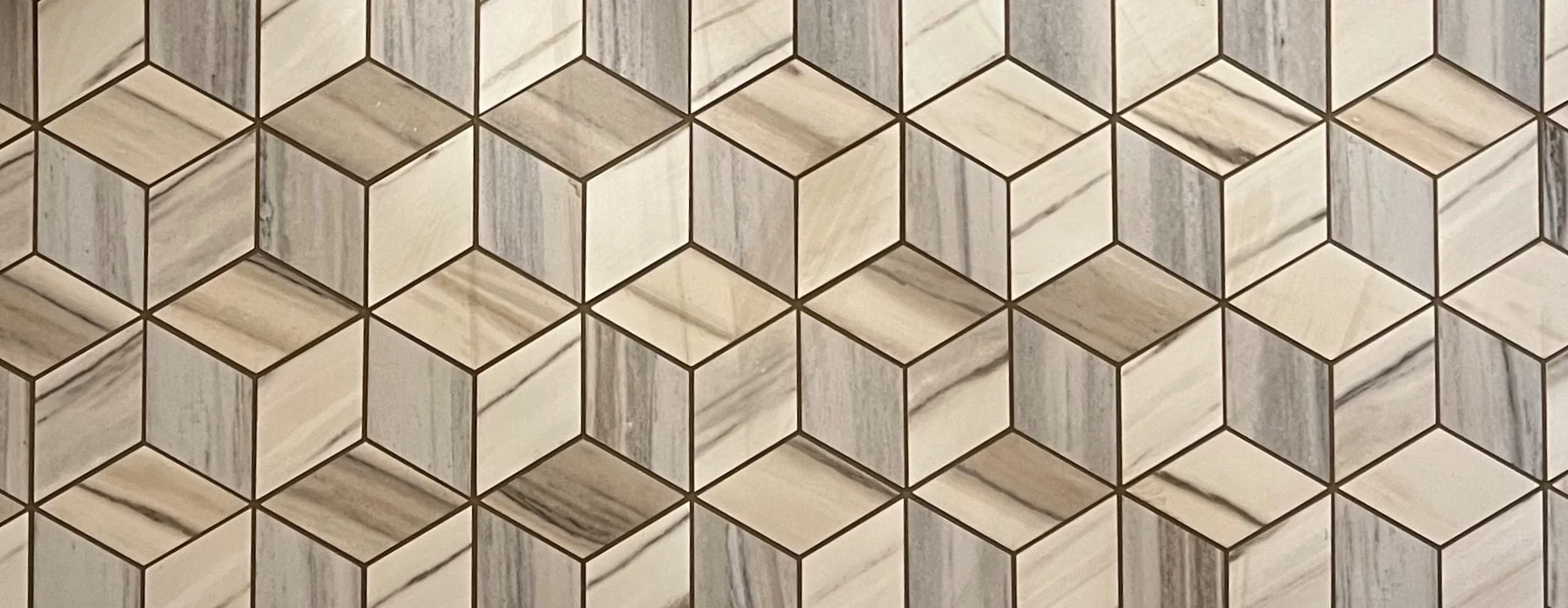 Close-up of a geometric tile wall featuring diamond shaped tile in a pattern of interconnected 3D cube shapes in shades of white, gray, and beige.