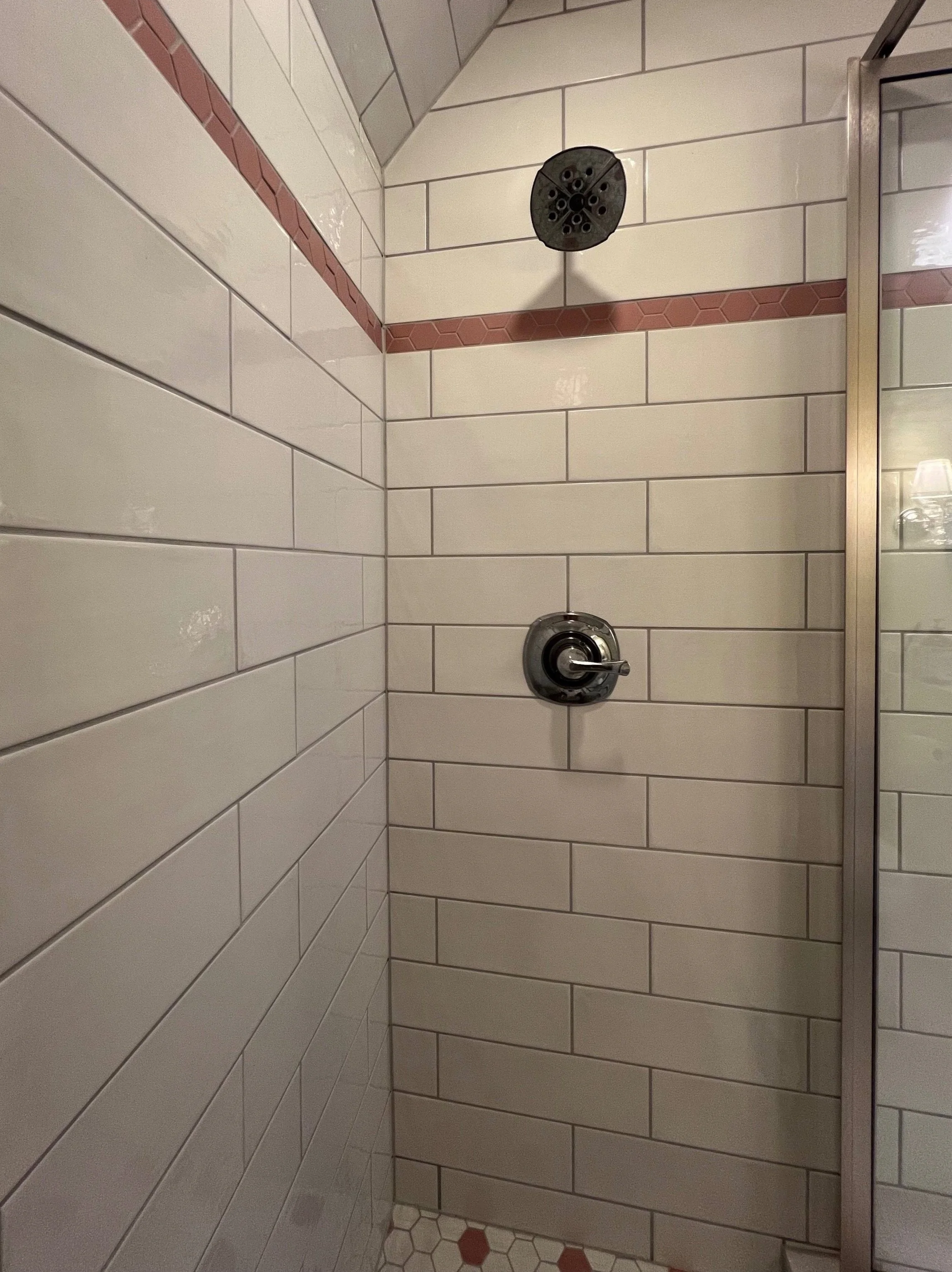 A shower wall with white rectangular tiles and a strip of small hexagonal tiles in brown and red. There is a black shower head at the top and a silver faucet below it. To the right, a glass shower partition with metal frame is visible.