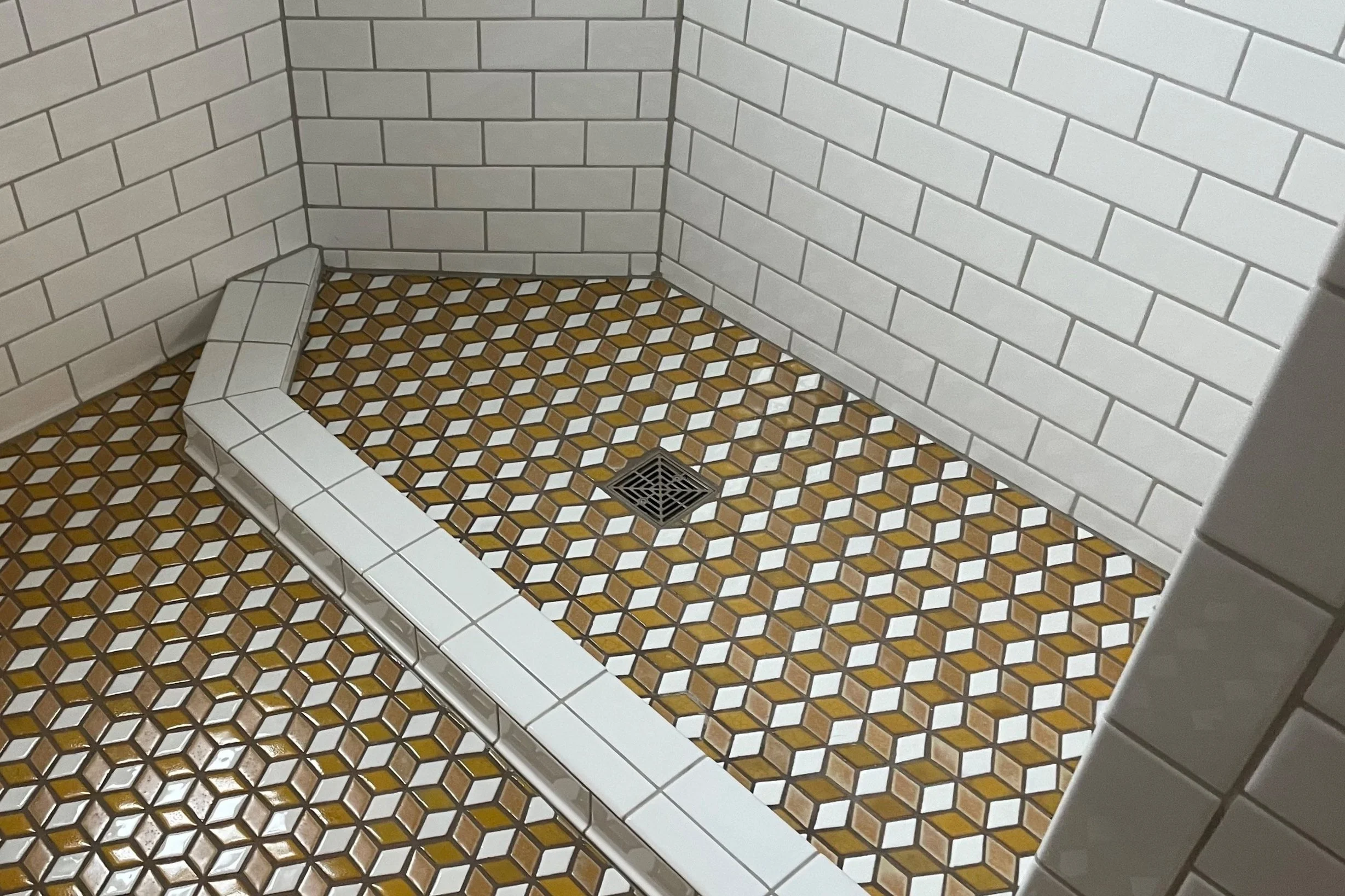 A view of a custom shower with white subway tiles on the walls and geometric-patterned floor tiles in shades of brown, white, and yellow. A small square drain is visible on the floor.