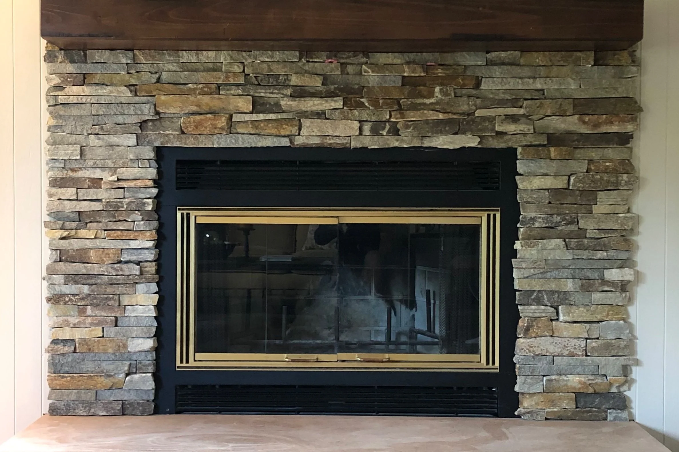 A fireplace surrounded with stacked stone elements and a wooden mantle on top and gold-colored trim around the fireplace opening.