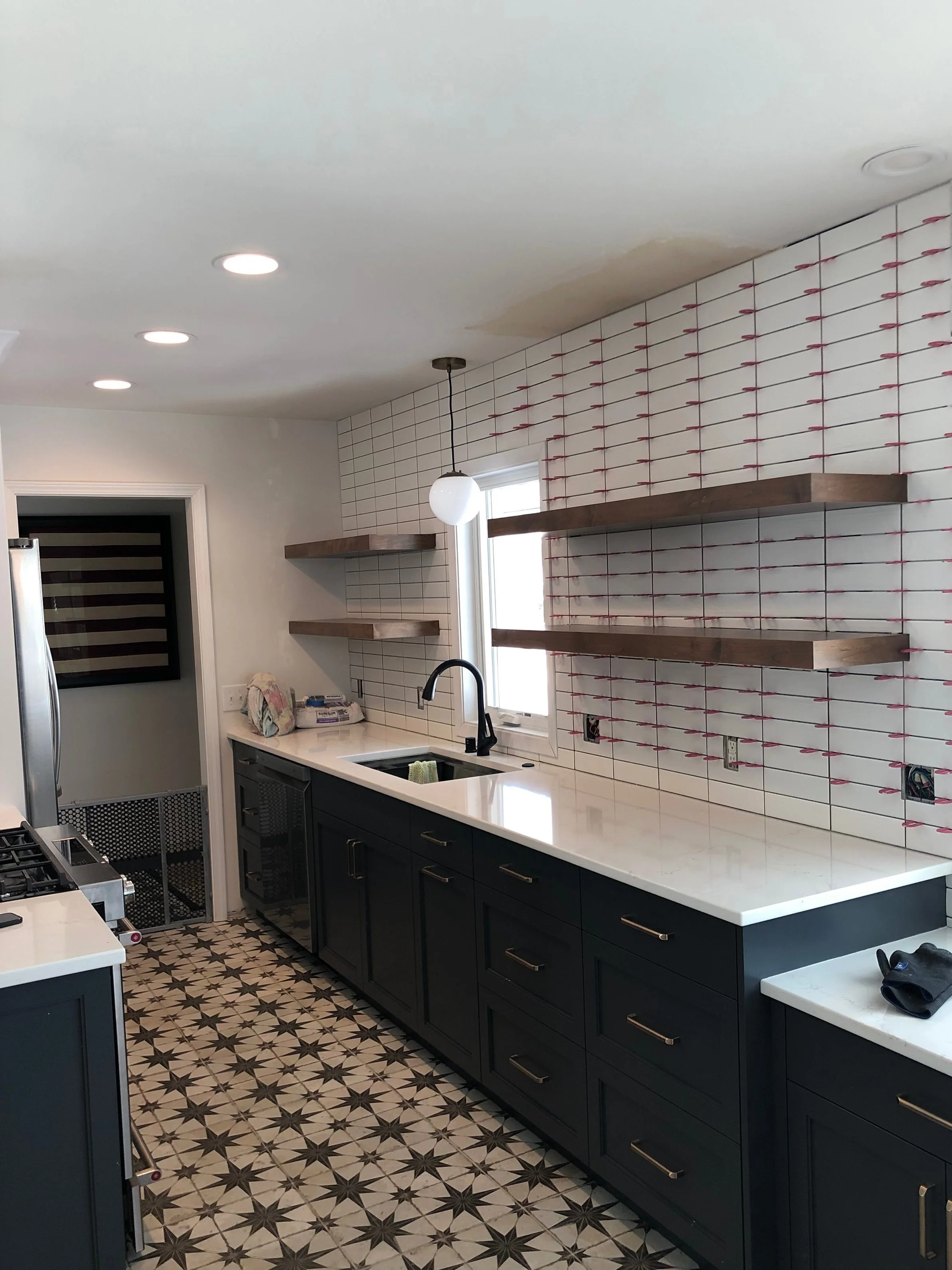 A kitchen with dark blue cabinets, white countertops, and patterned tile flooring. The walls are covered with white subway tiles and floating shelves on either side of a window set above the sink.