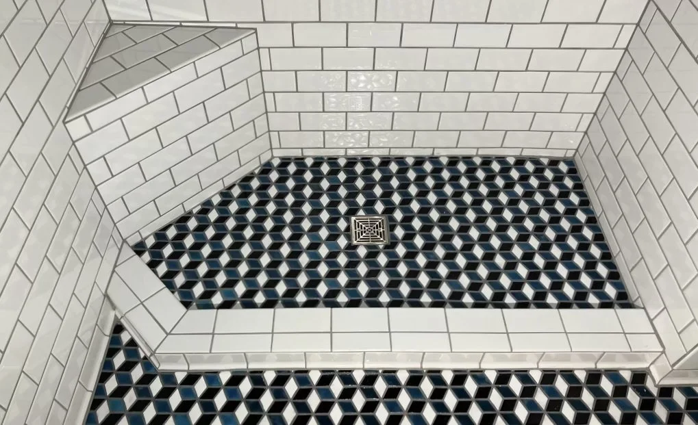 A custom shower with white subway tile on the walls and a geometric patterned floor in dark blue, black and white, a square drain is seen in the center of the shower pan with a small bench in the corner.