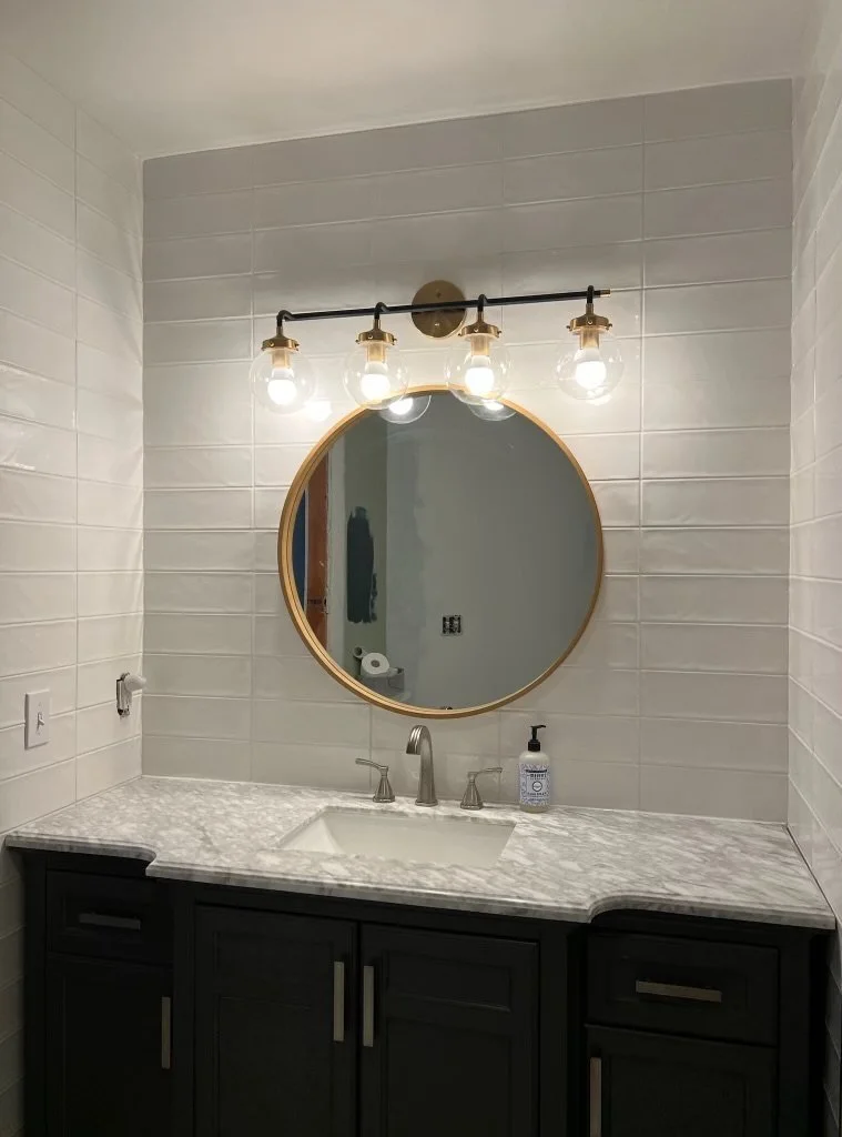 Bathroom vanity with white stacked subway tile, a round mirror, dark cabinet, marble countertop, faucet, and four exposed light bulbs above the mirror.
