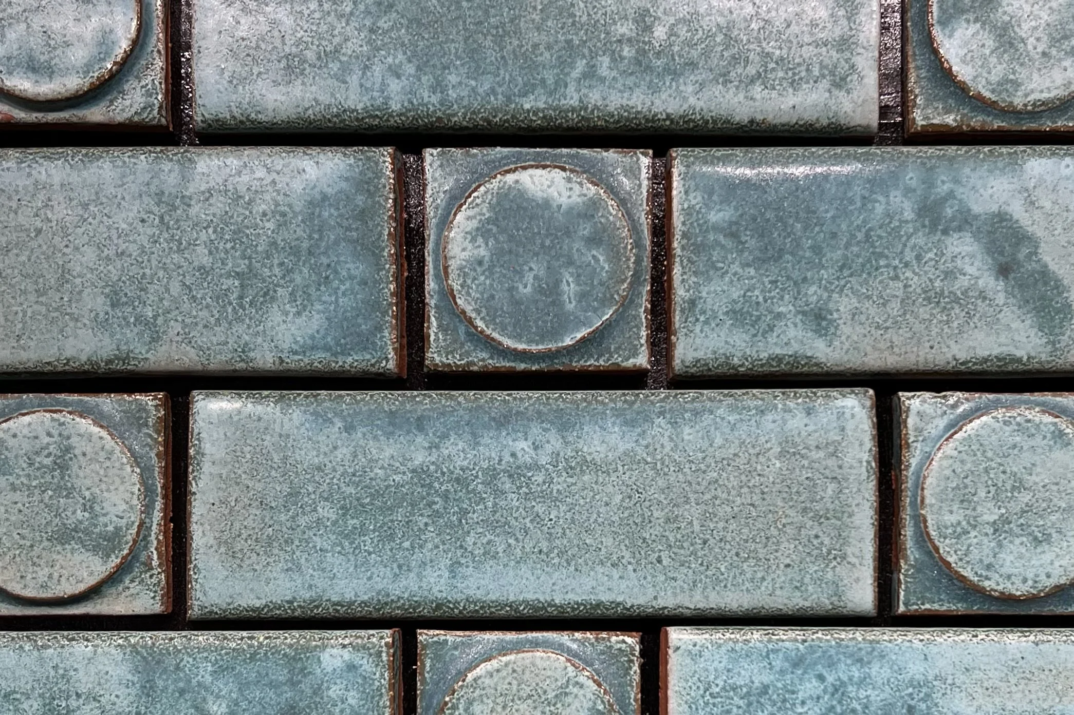 Close-up view of a section of textured green ceramic tiles with rounded and circular patterns. The tiles show signs of aging and rust stains along the edges.