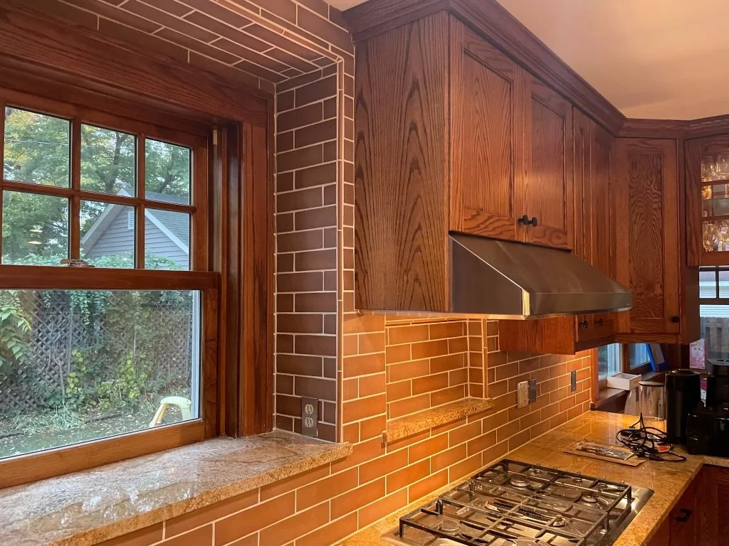 A kitchen backsplash with wooden cabinets, subway backsplash, a gas stove, and a window overlooking a backyard.