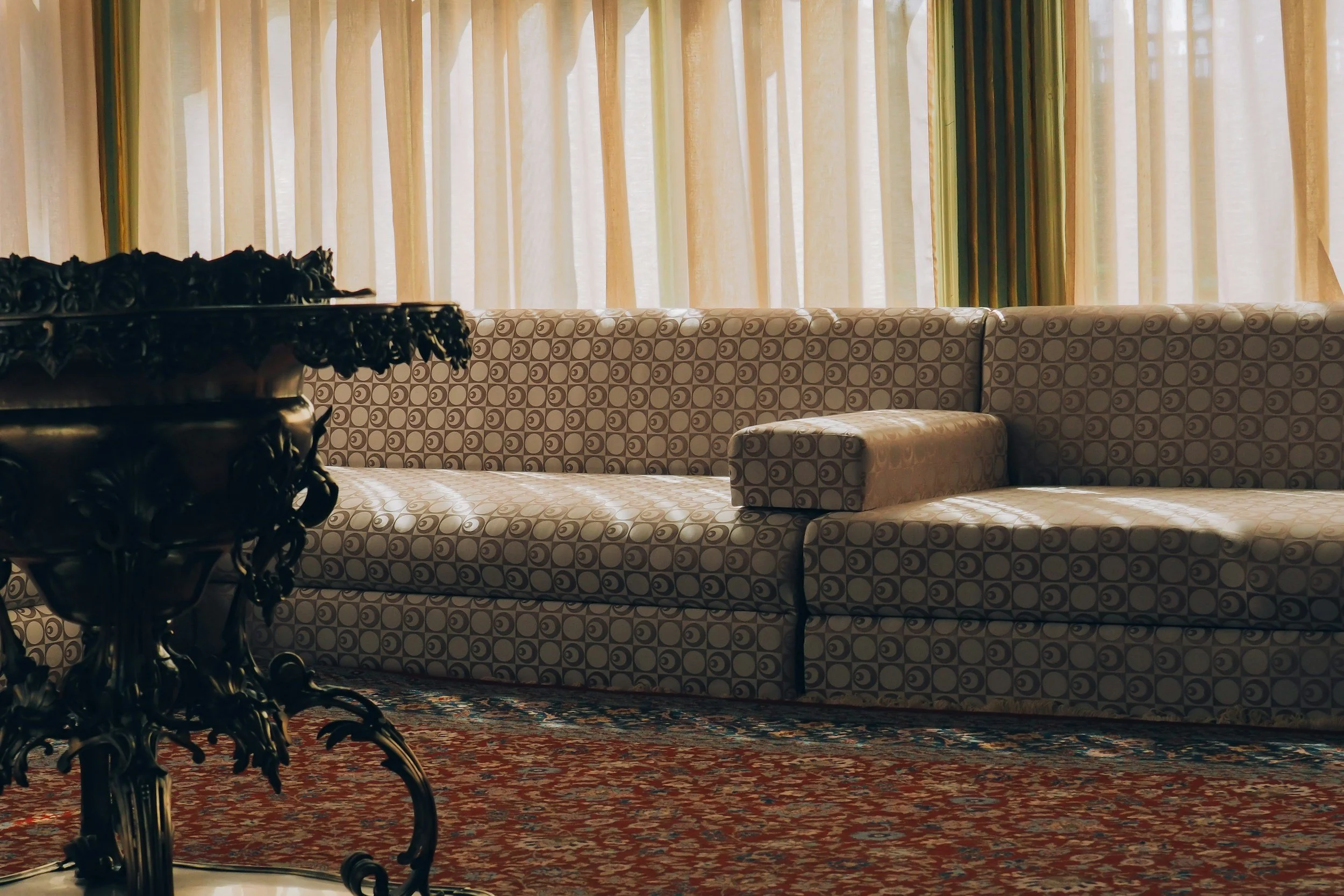 A vintage-style room with a large patterned sofa, ornate dark wooden table, and patterned curtains.