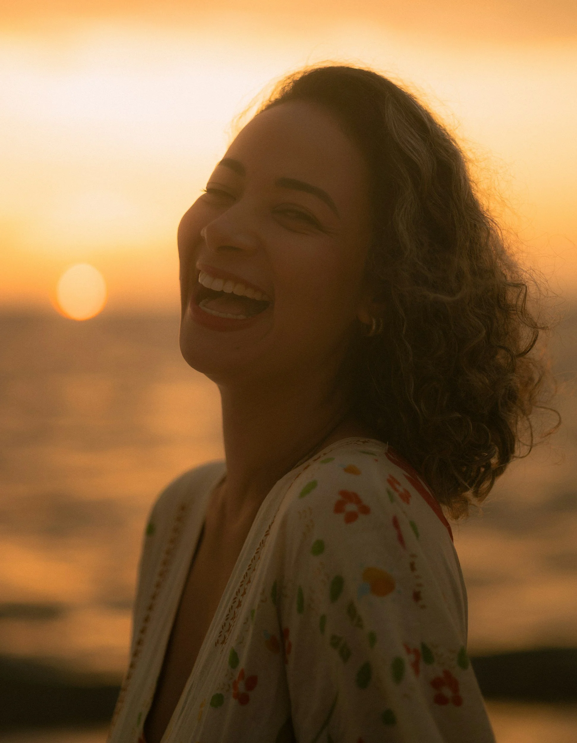 A woman with curly hair laughing at sunset on the beach.