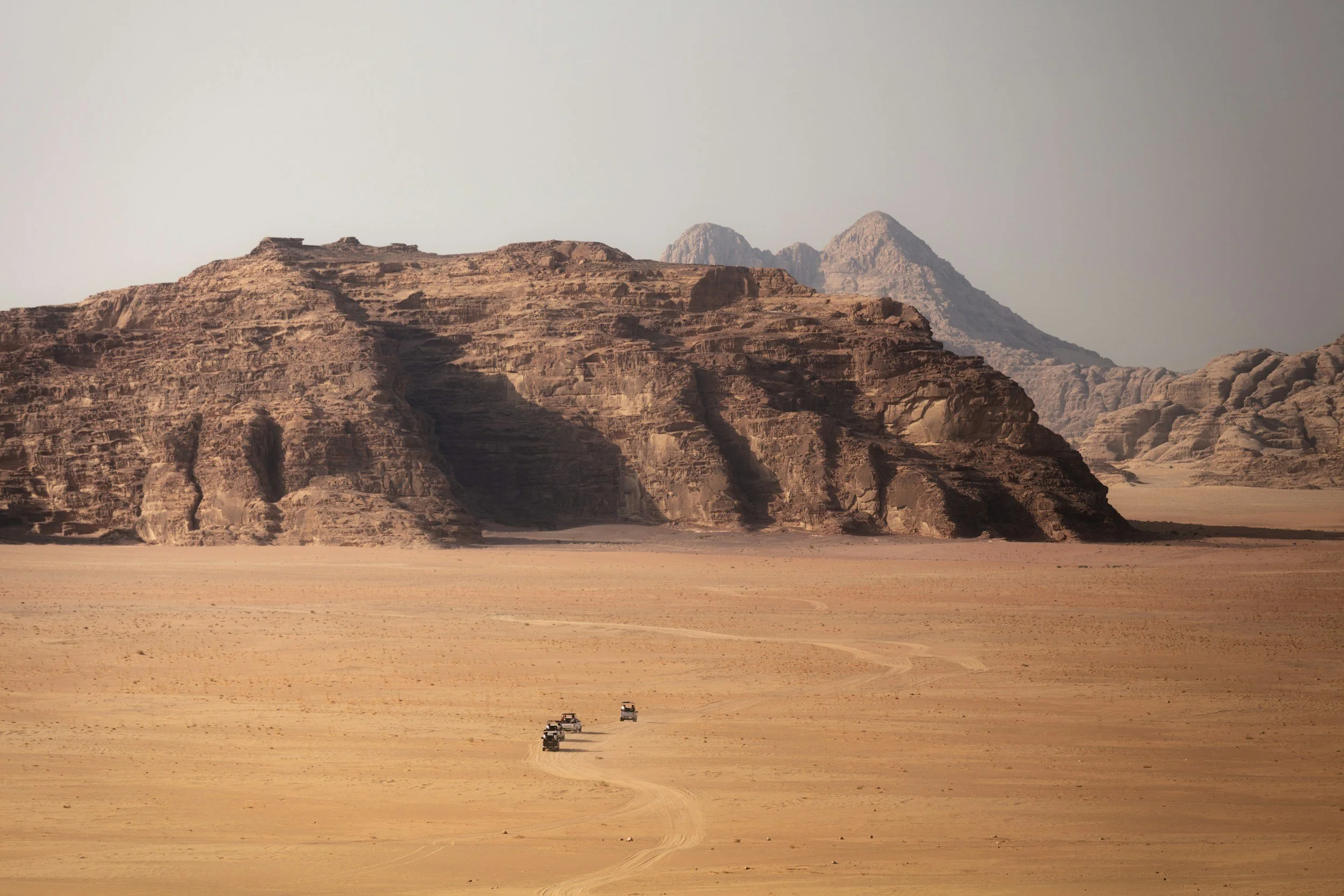 View of a desert with large rocky formations in the distance and a few vehicles driving along a path in the sand.