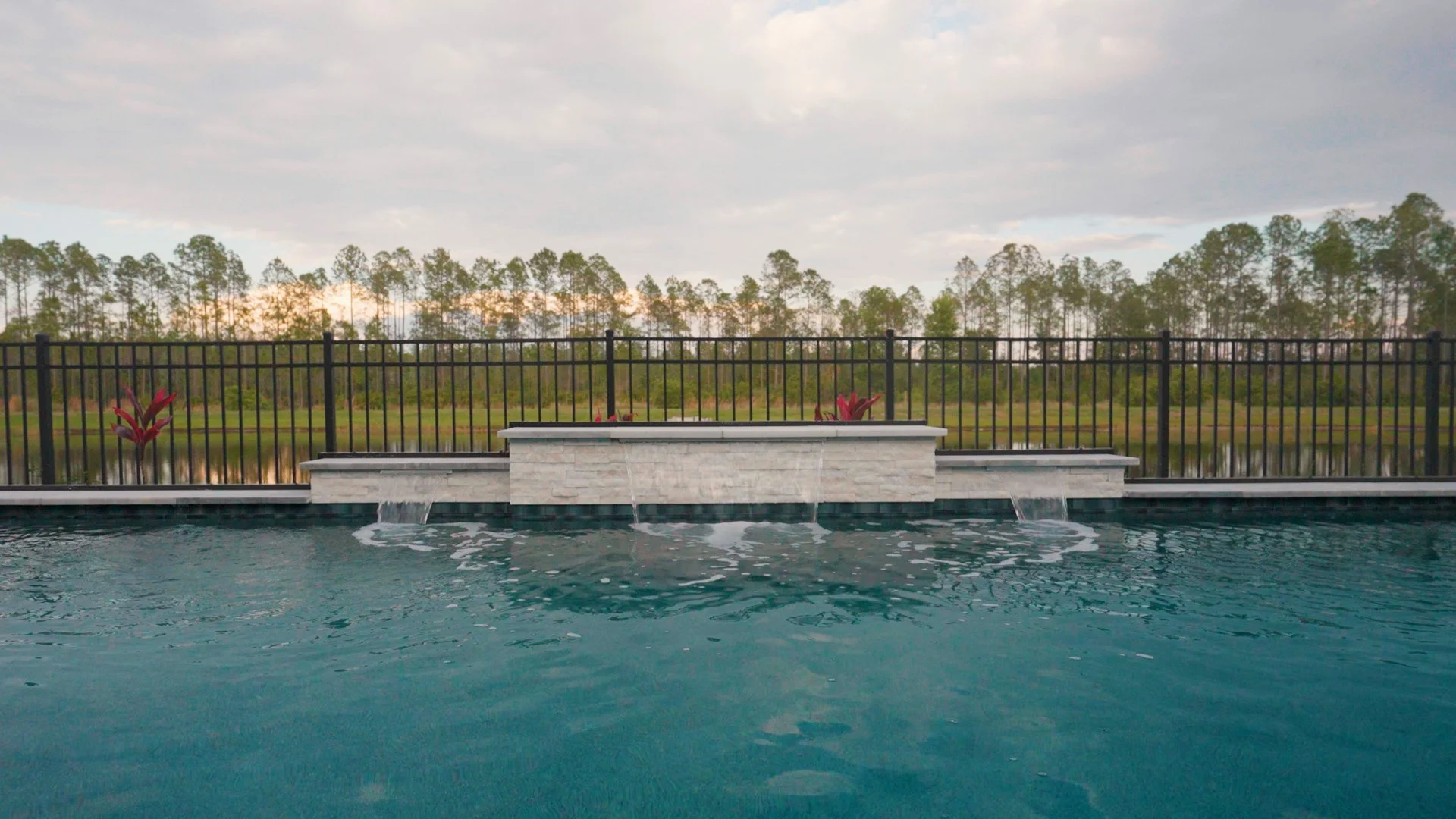 A swimming pool overlooking a natural landscape with trees and water. The pool has three small waterfalls flowing into it, bordered by a stone ledge and a black metal fence. Two red plants are placed on each side of the waterfalls.