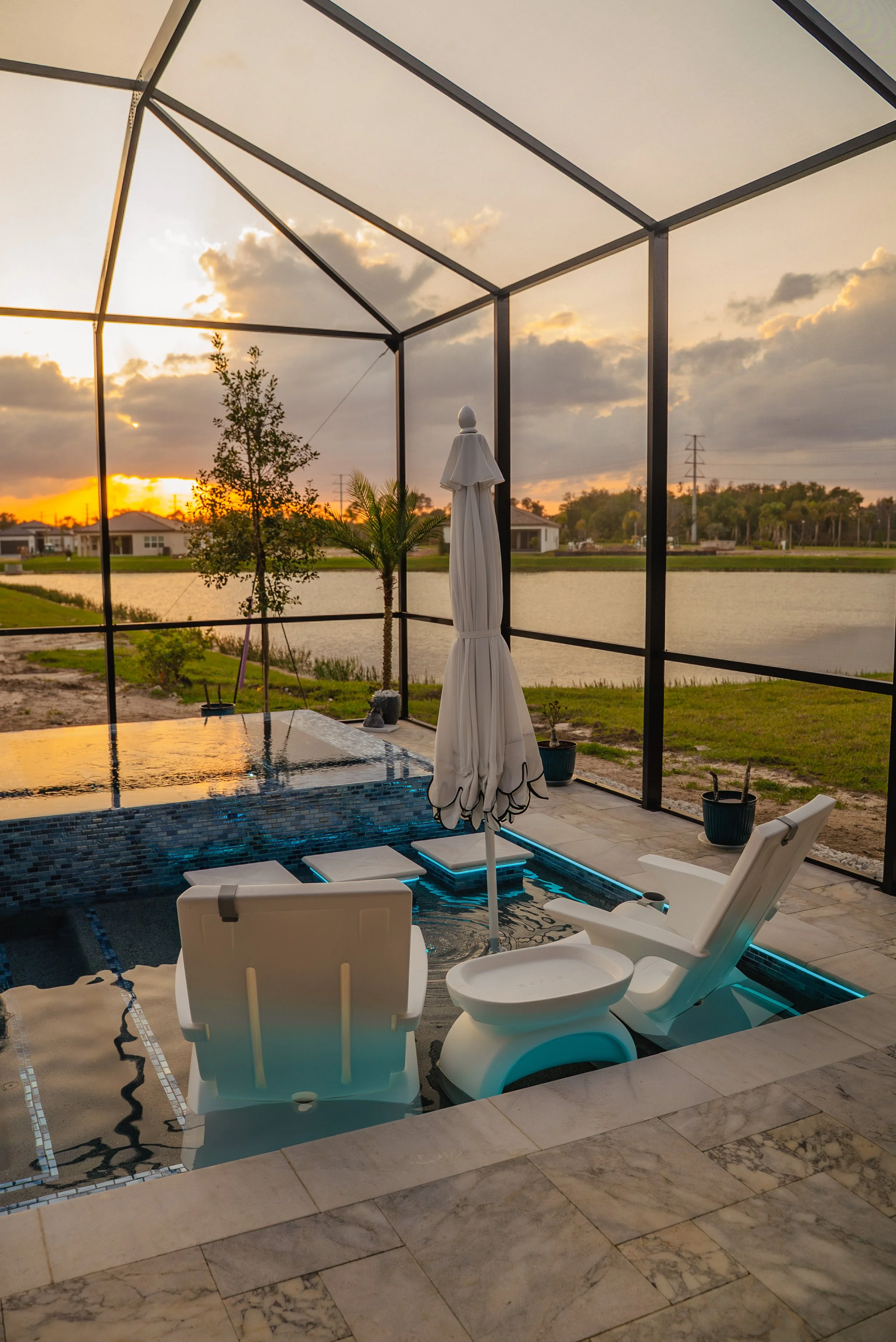 A covered patio with a view of a lake and houses in the background during sunset. There are pool chairs, an umbrella, a hot tub, and potted plants.
