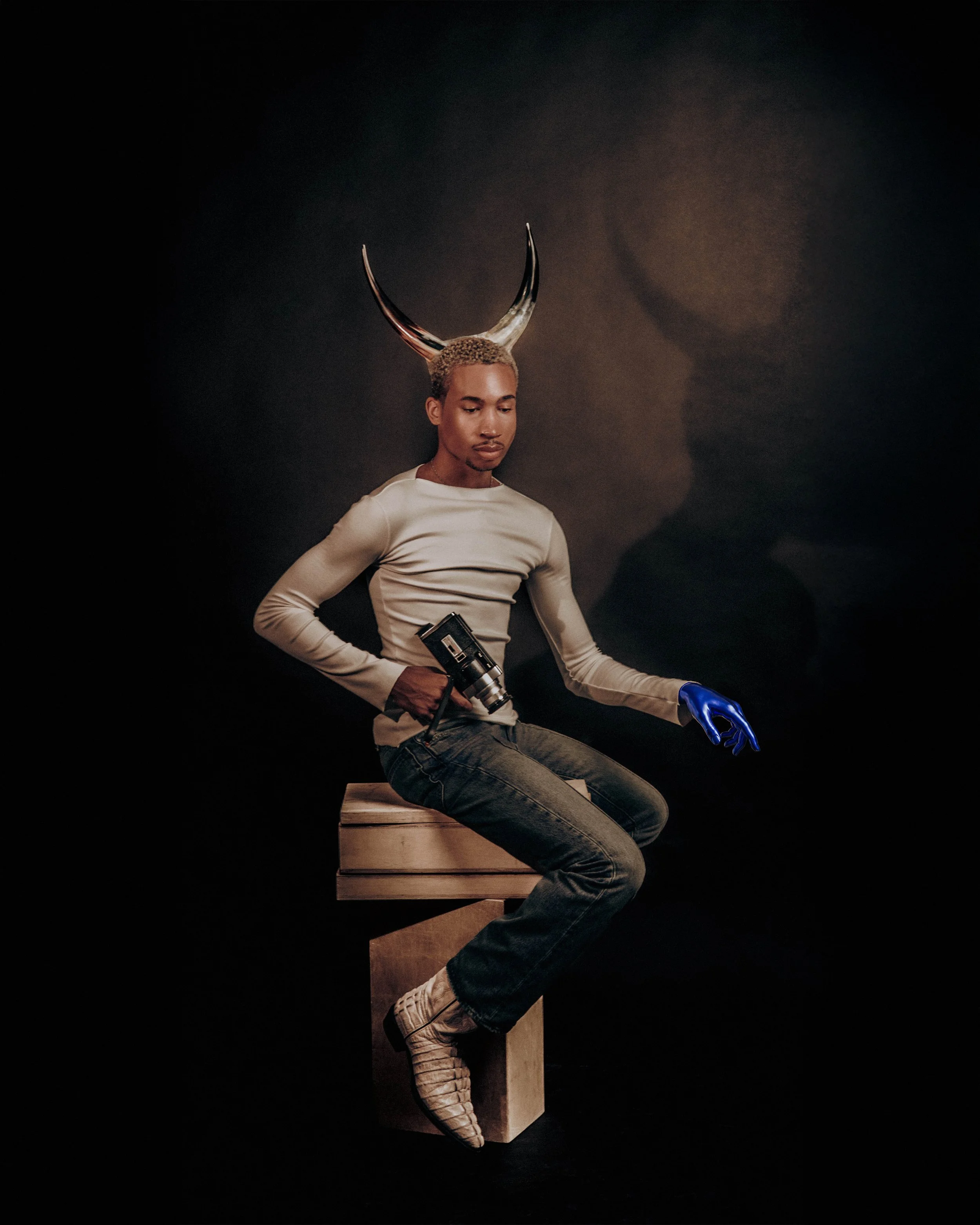 A person with light skin and short, curly blond hair is sitting on a wooden block against a black background. They are wearing a beige long-sleeve shirt, blue jeans, beige shoes, and a blue glove on their right hand. They are holding a vintage camera with their left hand and have a large set of horns on the person's head, which appear to be an artistic or costume piece.