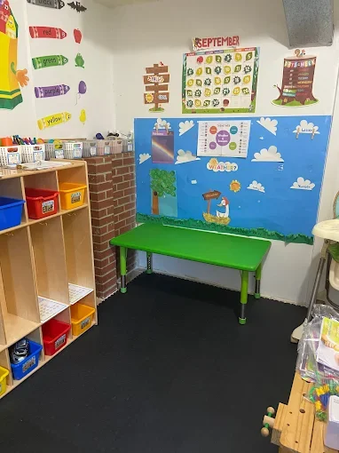 A classroom corner with a green bench, a colorful bulletin board with a whale and cloud scene, a calendar for September, and wall decorations including colors and shapes. There are storage bins on a wooden shelf and a small table with supplies.