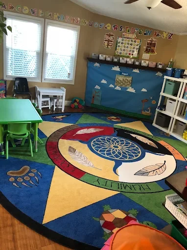 A colorful classroom corner with a large circular rug featuring a dreamcatcher design and feathers. There is a green plastic table with matching chairs, a wall decorated with an alphabet banner, and shelves with educational materials. Windows allow n