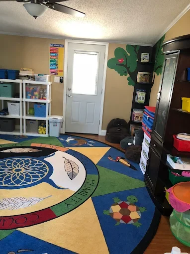 A colorful classroom corner with a door, storage shelves, a rug with animal and nature motifs, a green tree painted on the wall, and various educational supplies.