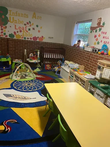 A preschool classroom with colorful decorations, a bright rug, play area with toys and a small table with chairs, a window, and wall art with text about infants.