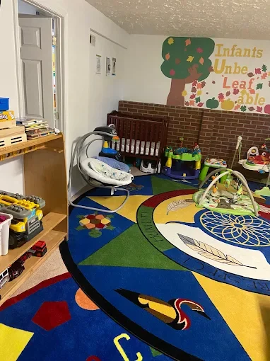 Children's playroom with colorful circular rug, baby rocker, toys, books, and wall decoration. The wall has a painted tree and the words 'Infants Unbe... Leaf ...able'.